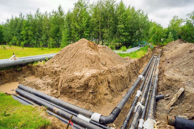 Black pipes are laid in a deep, excavated construction trench in a rural outdoor setting next to a large pile of dirt.