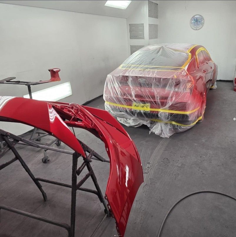 Red car in a paint booth, covered in plastic, with a red bumper.