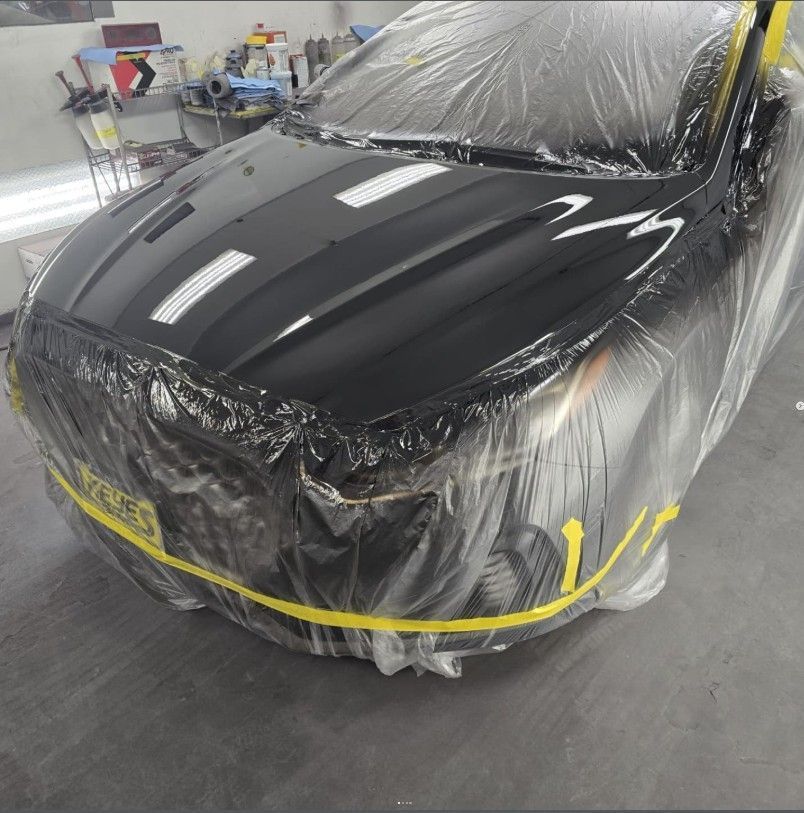 Black car covered in plastic sheeting and yellow tape in a repair shop.