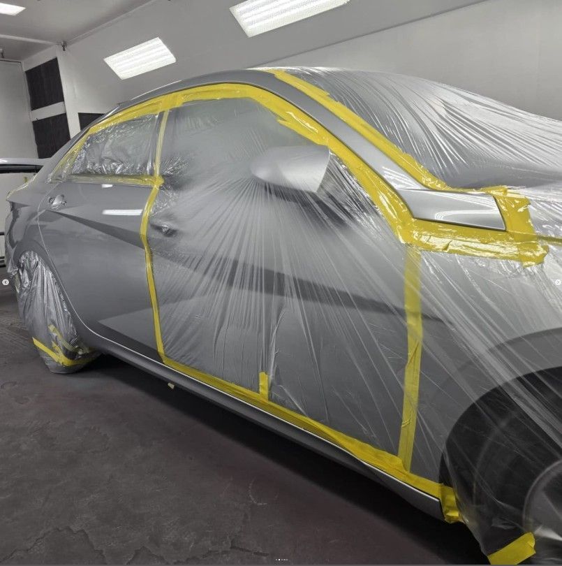 Silver car covered in plastic sheeting and yellow tape in a spray booth.