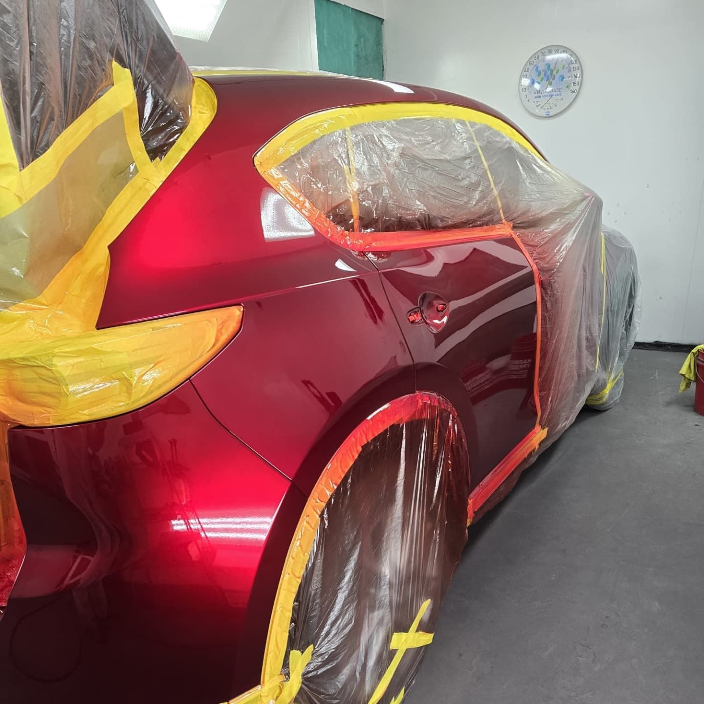 Red car partially covered in plastic and tape for painting in a workshop.