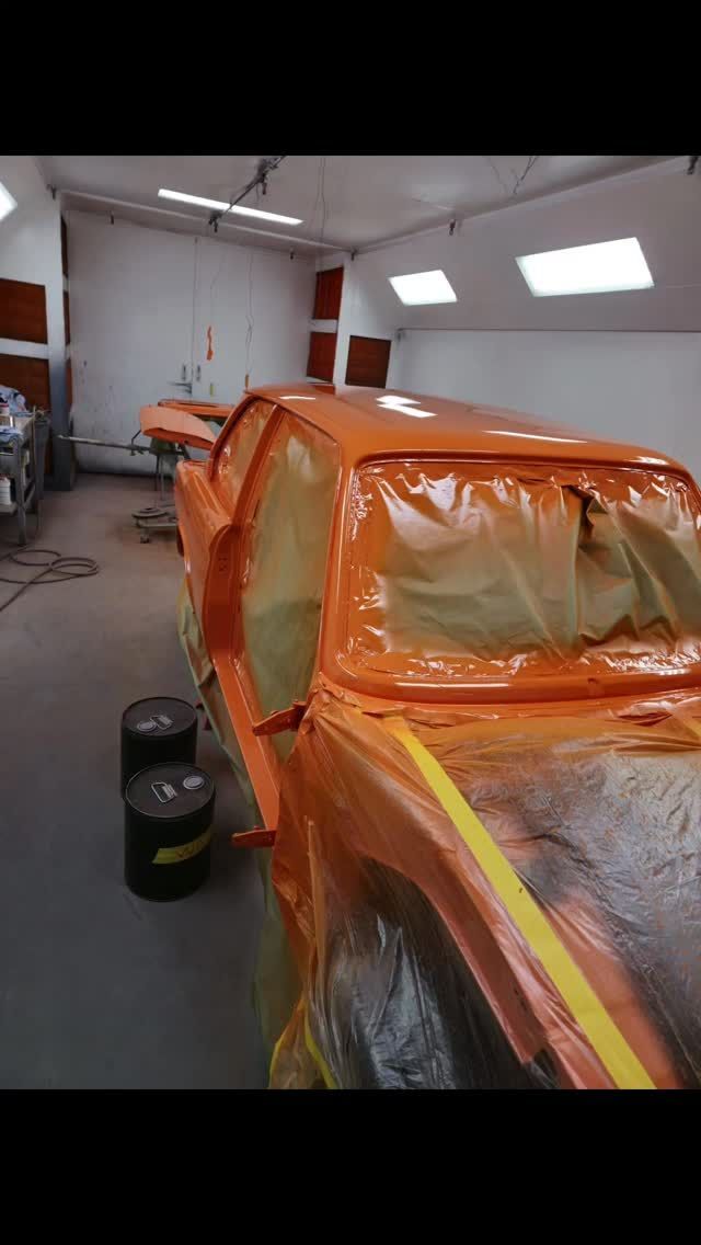 Orange car being painted in a paint booth, covered in plastic, with paint cans on the floor.