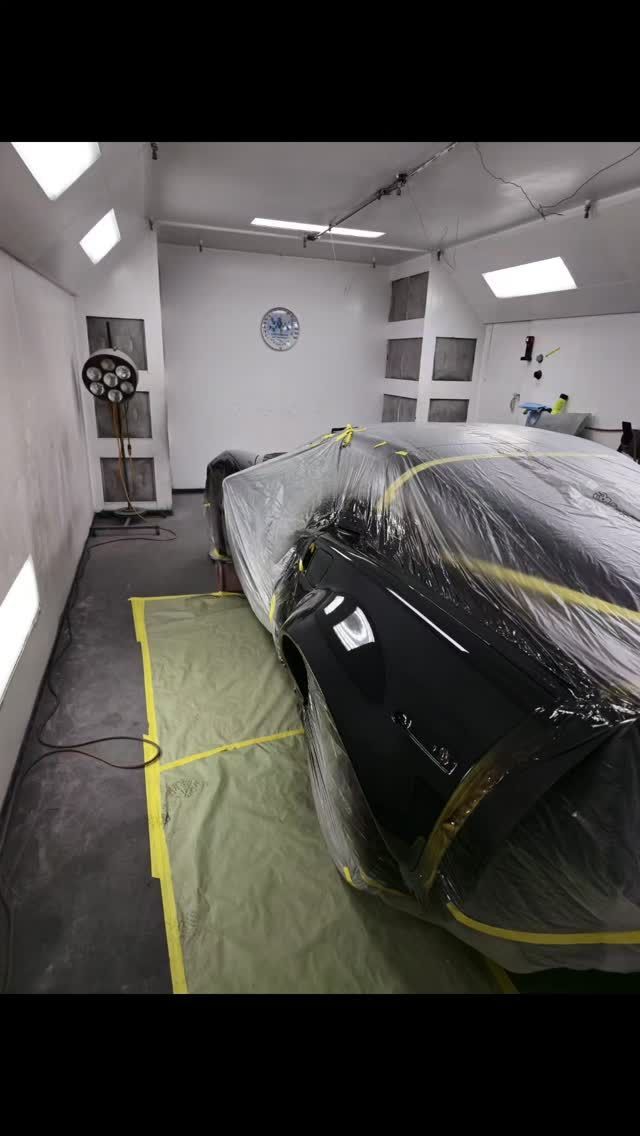Car in a paint booth, covered in plastic, on a green drop cloth. Spray booth with white walls and overhead lights.