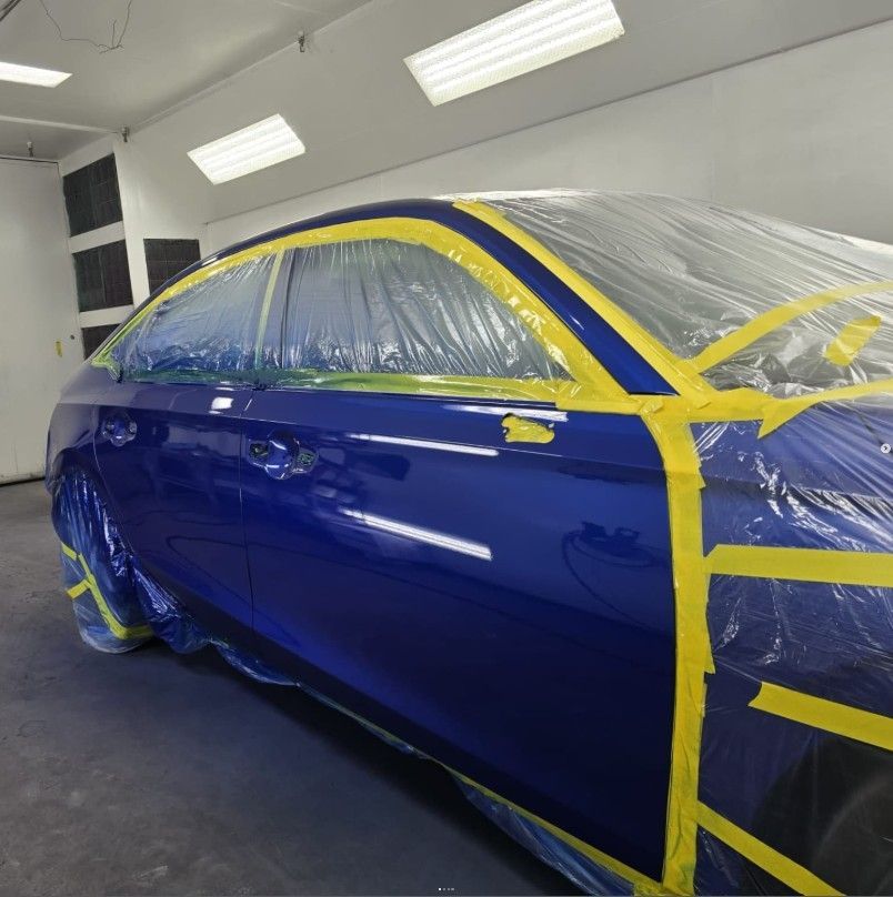 Blue car in a paint booth, taped with yellow tape and covered in plastic sheeting.
