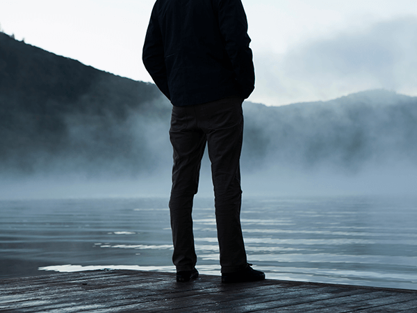 Person standing on a dock beside a misty lake, facing mountains at dawn