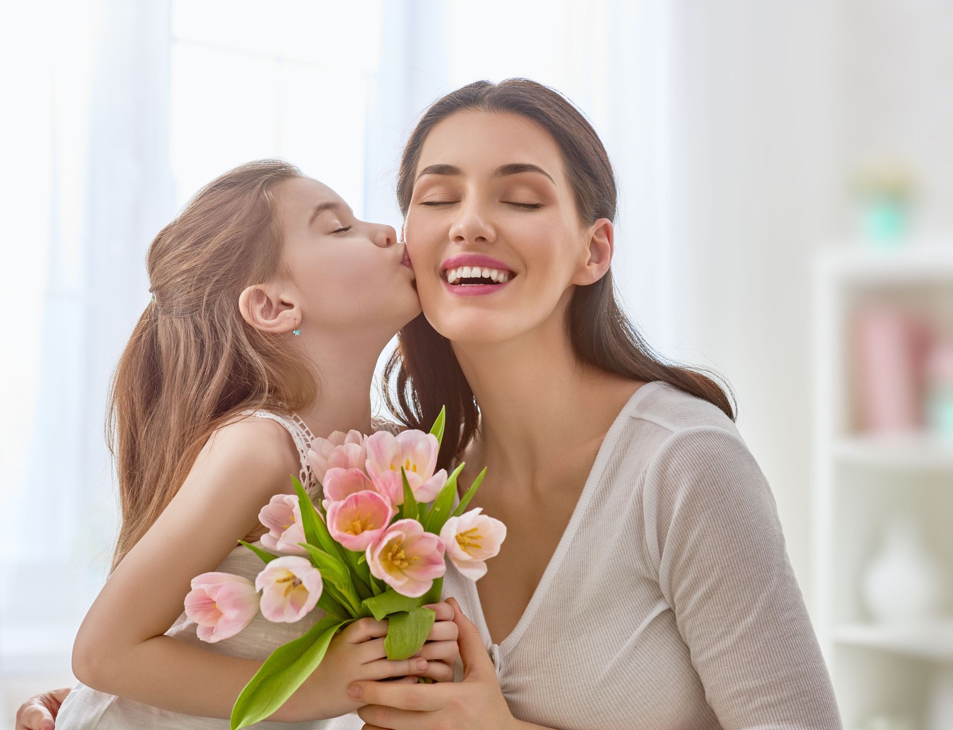 Child kissing smiling woman holding pink tulips indoors