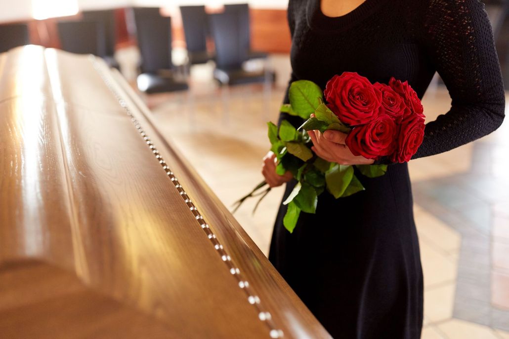 Person in black dress holding a bouquet of red roses beside a polished wooden casket