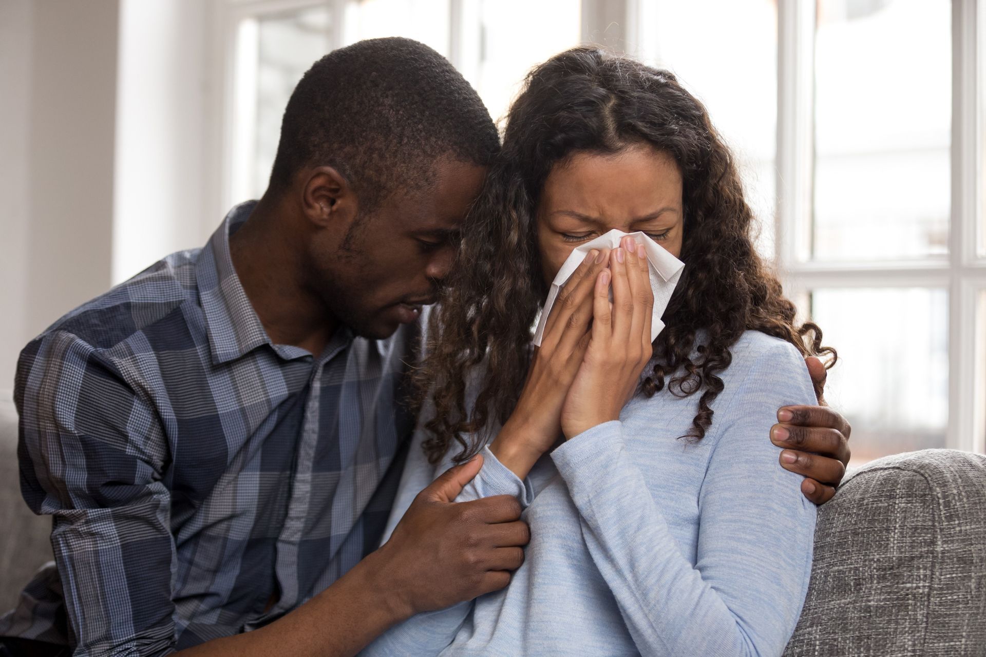 Couple on a couch, one comforting the other who is crying into a tissue.