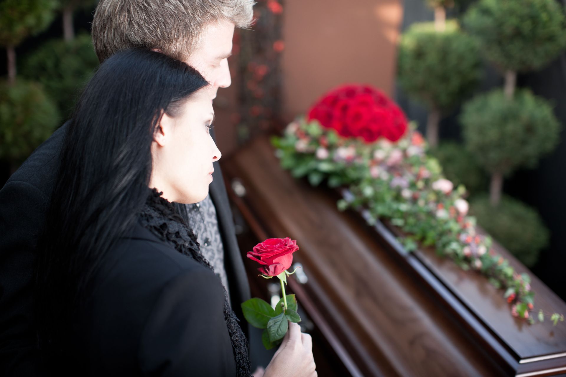 Two people place a red rose on a casket at a funeral