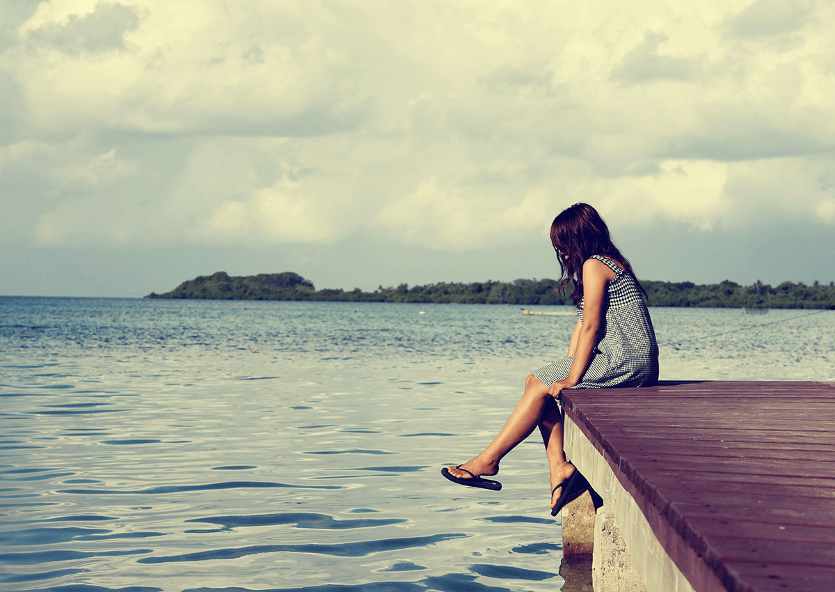Person sitting on a dock over calm water, looking toward a distant island at sunset