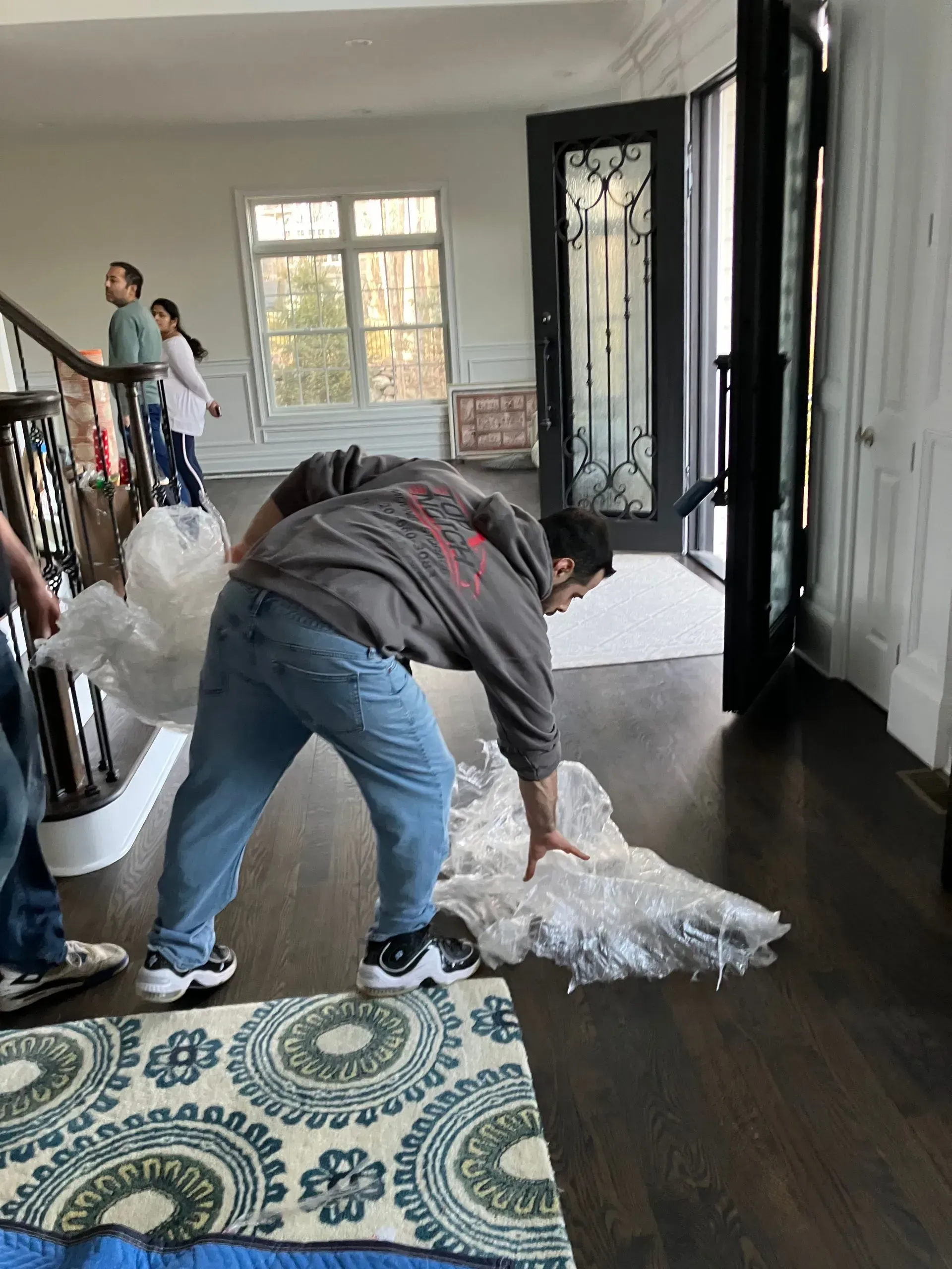 Person unpacking bubble wrap on dark wood floor, another person and children in the background.