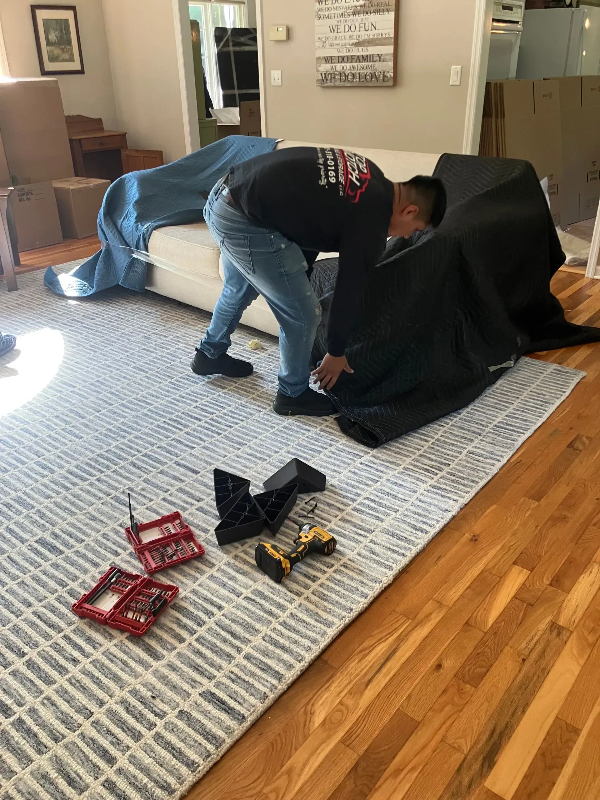 Man installing furniture, tools on rug. Sofa and chair covered, hardwood floor, natural light.
