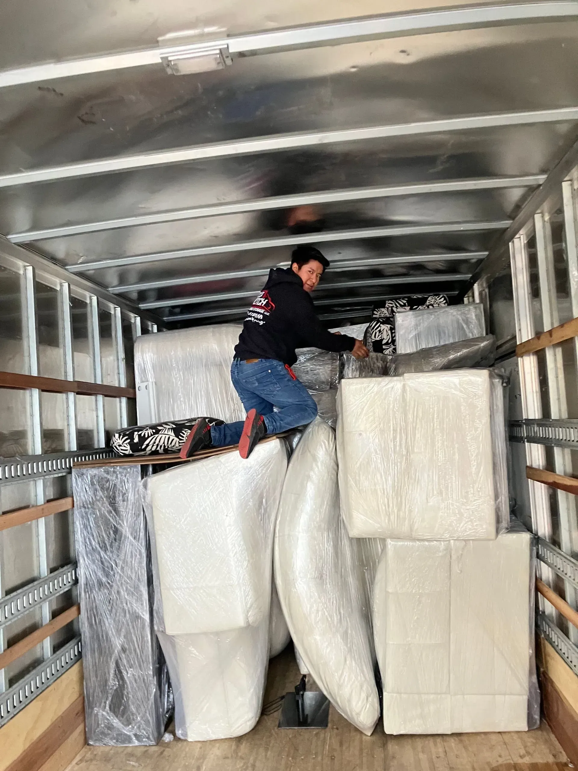 A person in a moving truck, surrounded by wrapped furniture, reaching for an item.