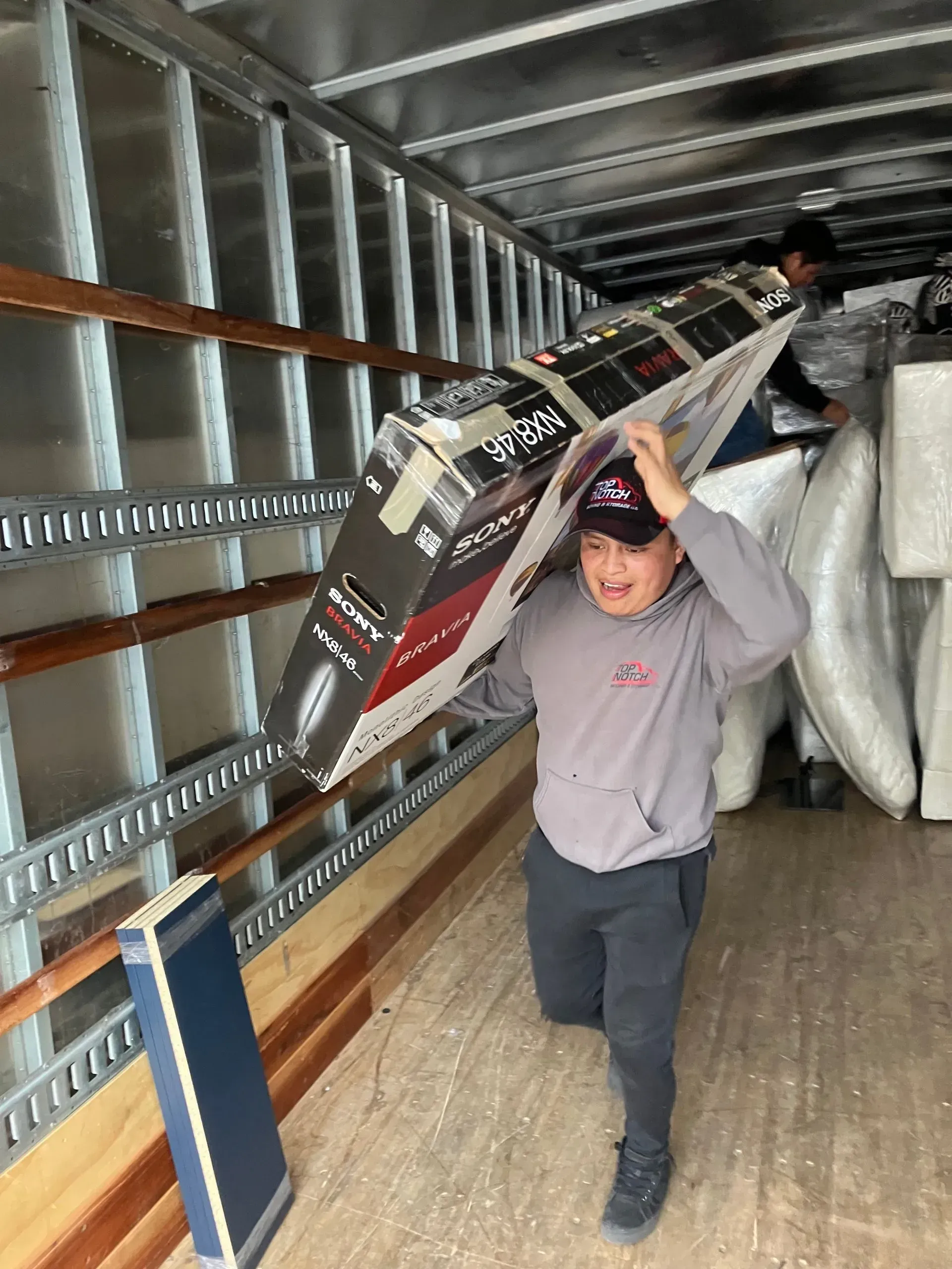 Person carrying a large TV box inside a moving truck. Another person in background.