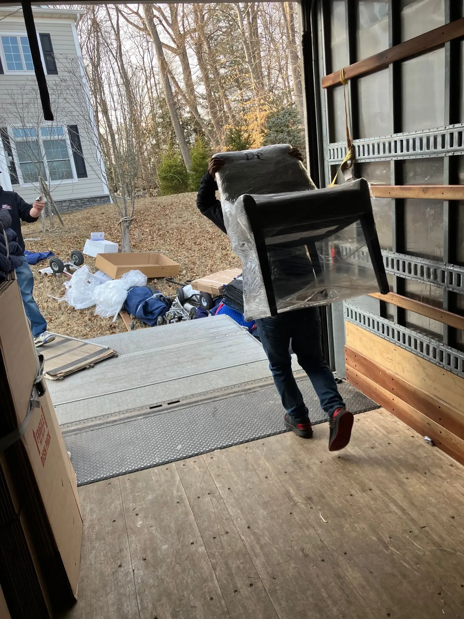 Person carrying a chair wrapped in plastic into a moving truck. Boxes and other items are outside.