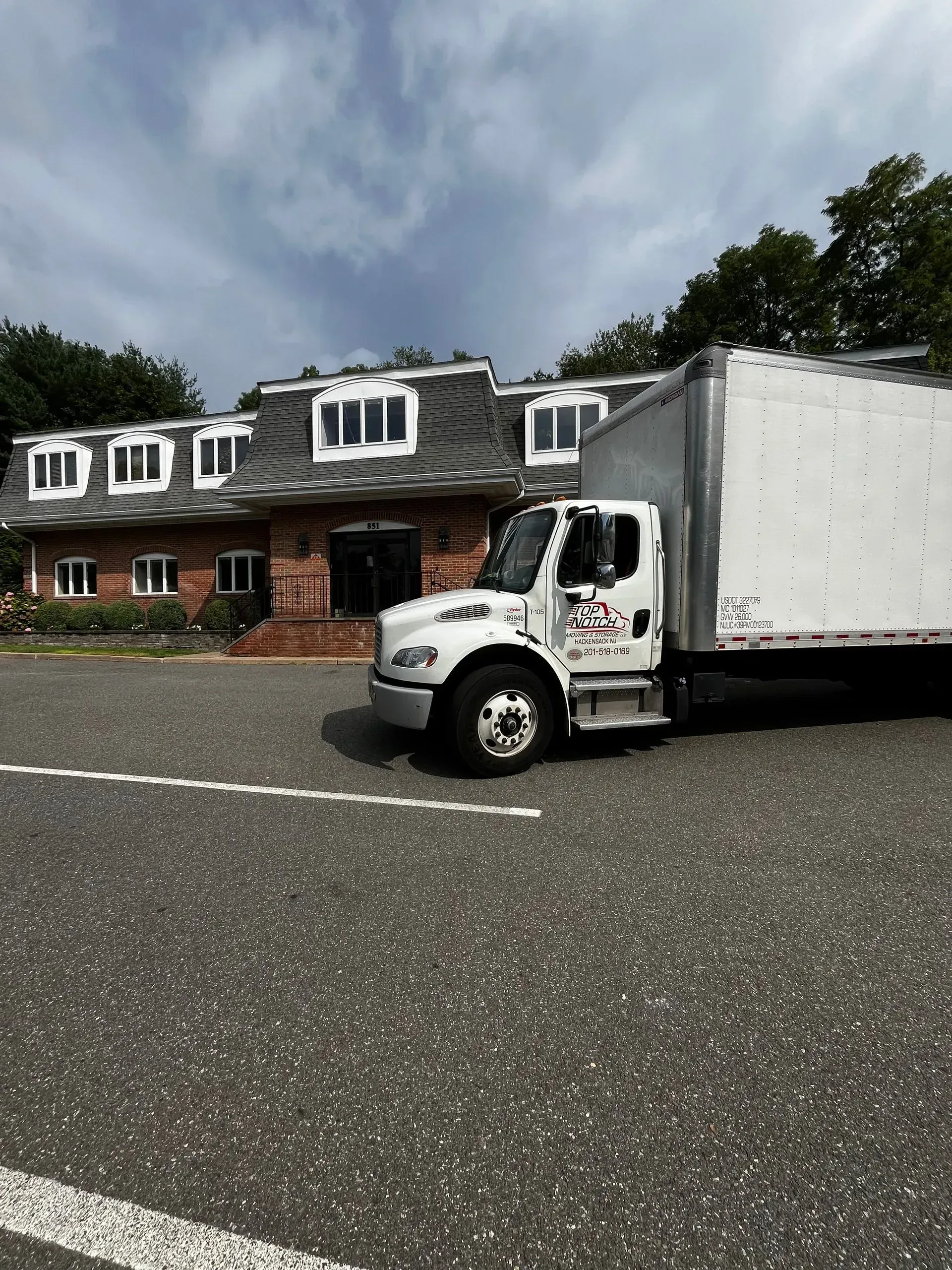 White box truck parked in front of a brick building with a gray roof and white windows under a cloudy sky.