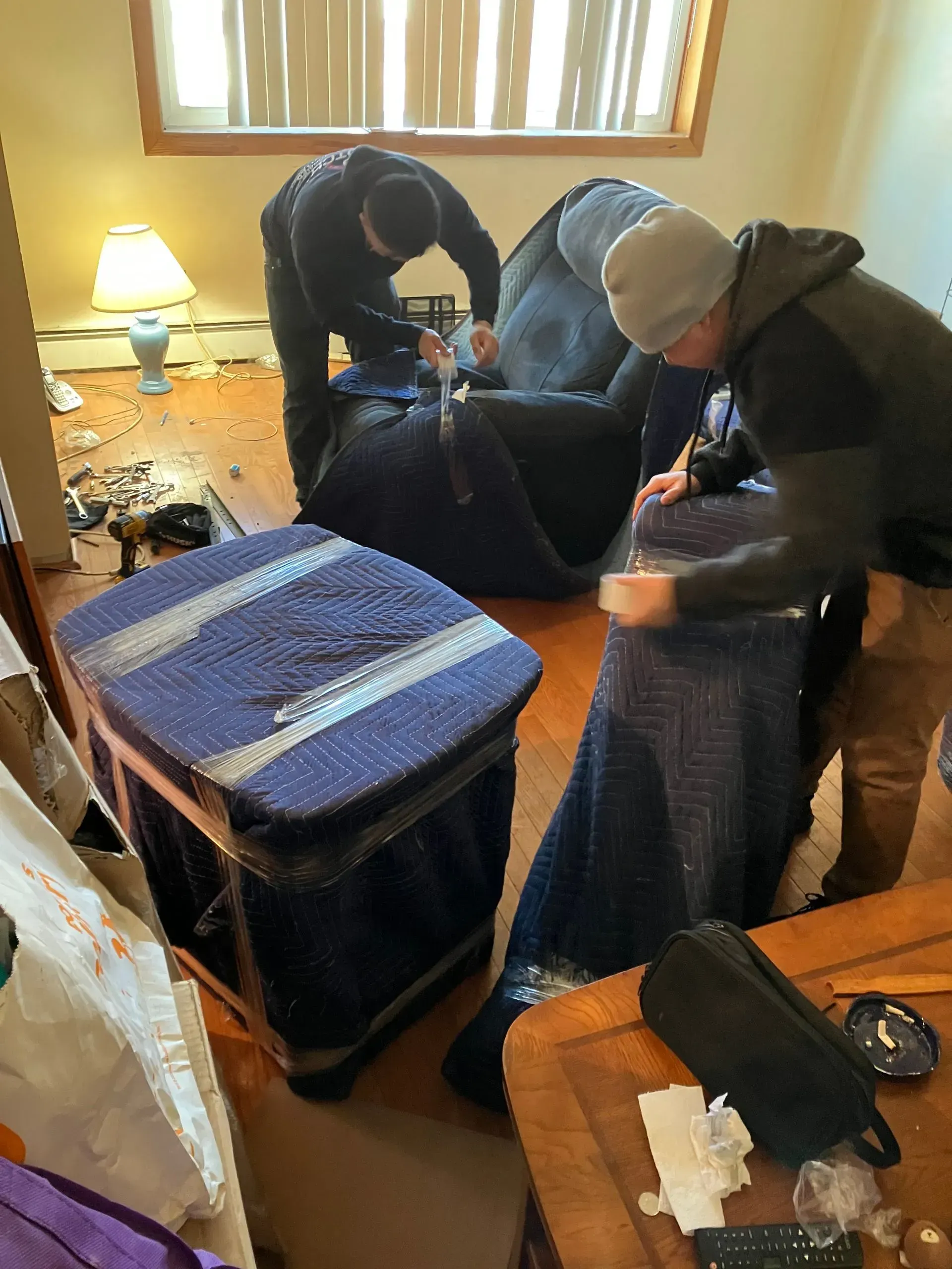 Three people packing furniture with blue padding in a room with a window and lamp.