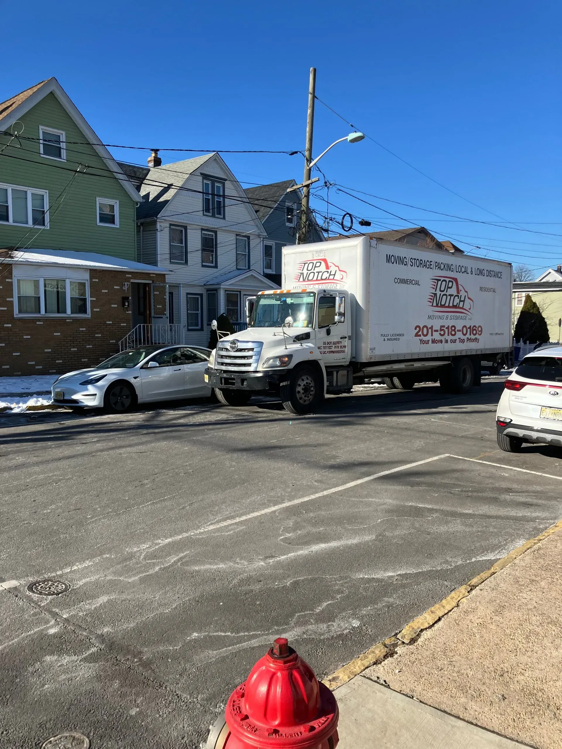 White moving truck parked on a street in front of houses; a car is parked in the foreground.