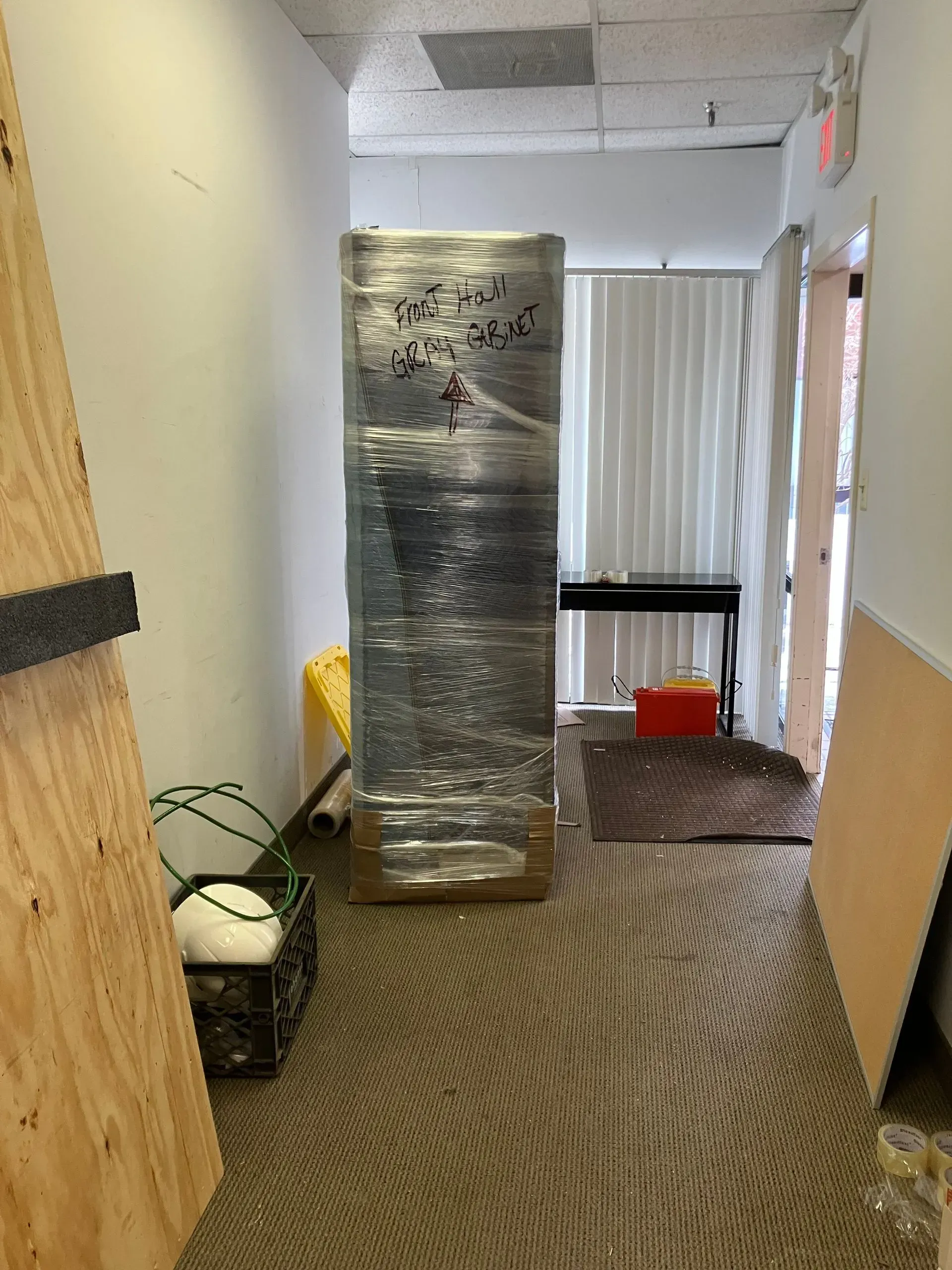 A tall, wrapped item stands in a narrow hallway with plywood and other items scattered around.