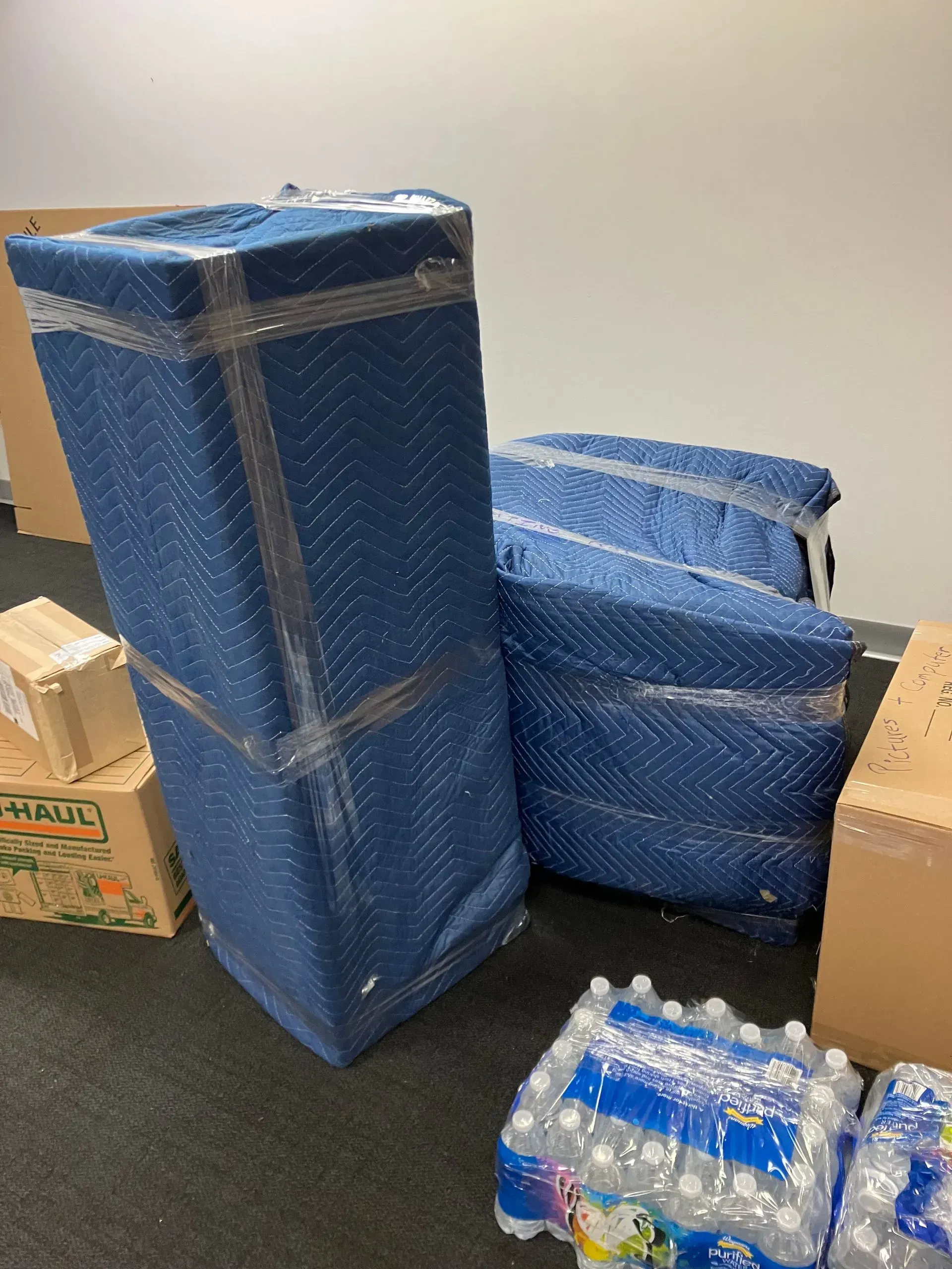 Moving items wrapped in blue padding and plastic wrap, water bottles, and boxes in a room.