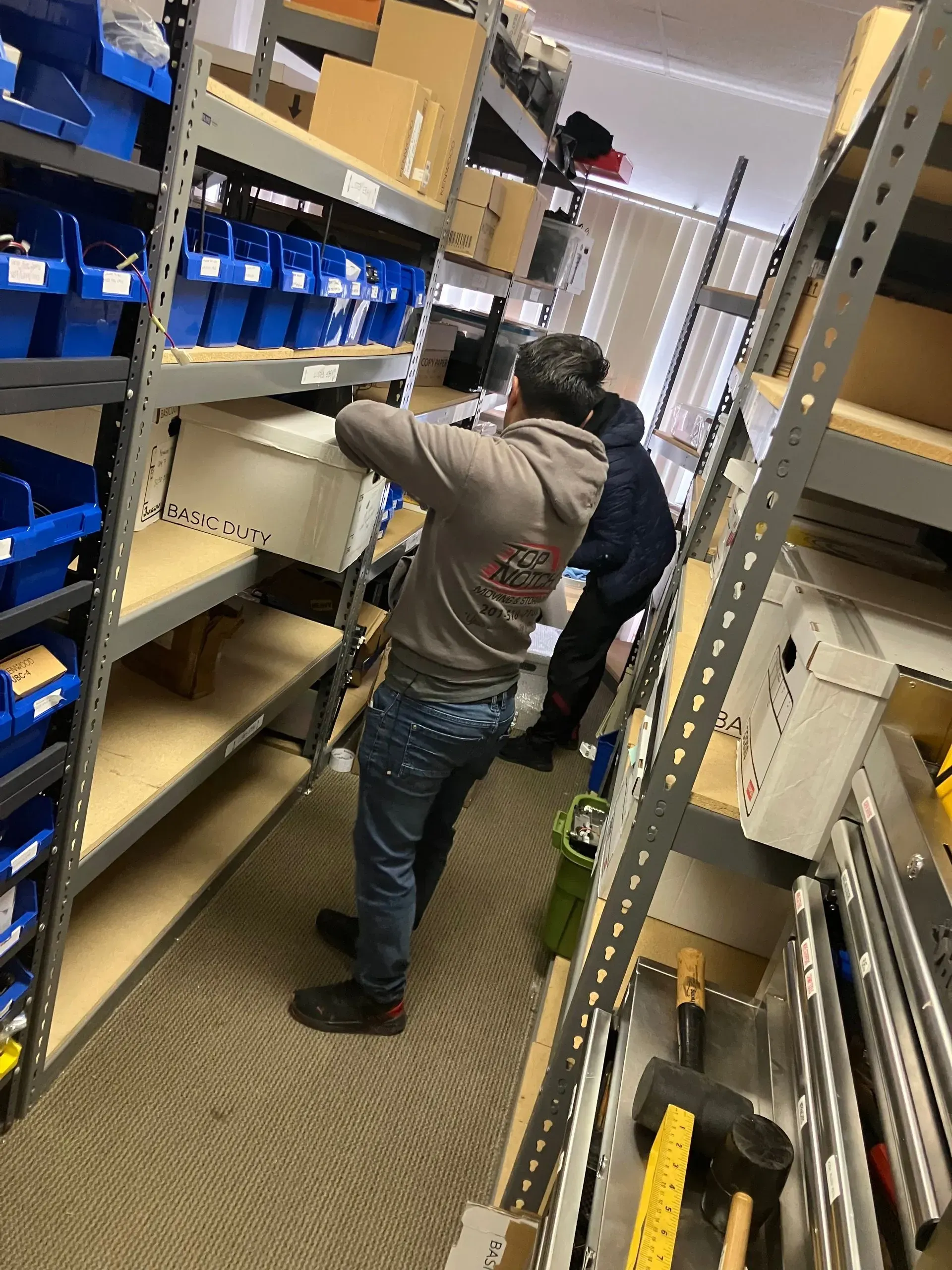 Two people in a storage room with shelves, removing a box. Blue bins, brown boxes, and tools present.