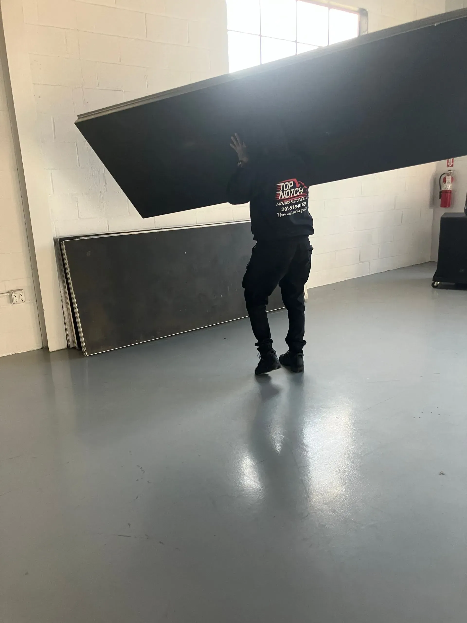 Person carrying large, dark rectangular panel in a room with gray floor and white walls. Another panel rests on the floor.