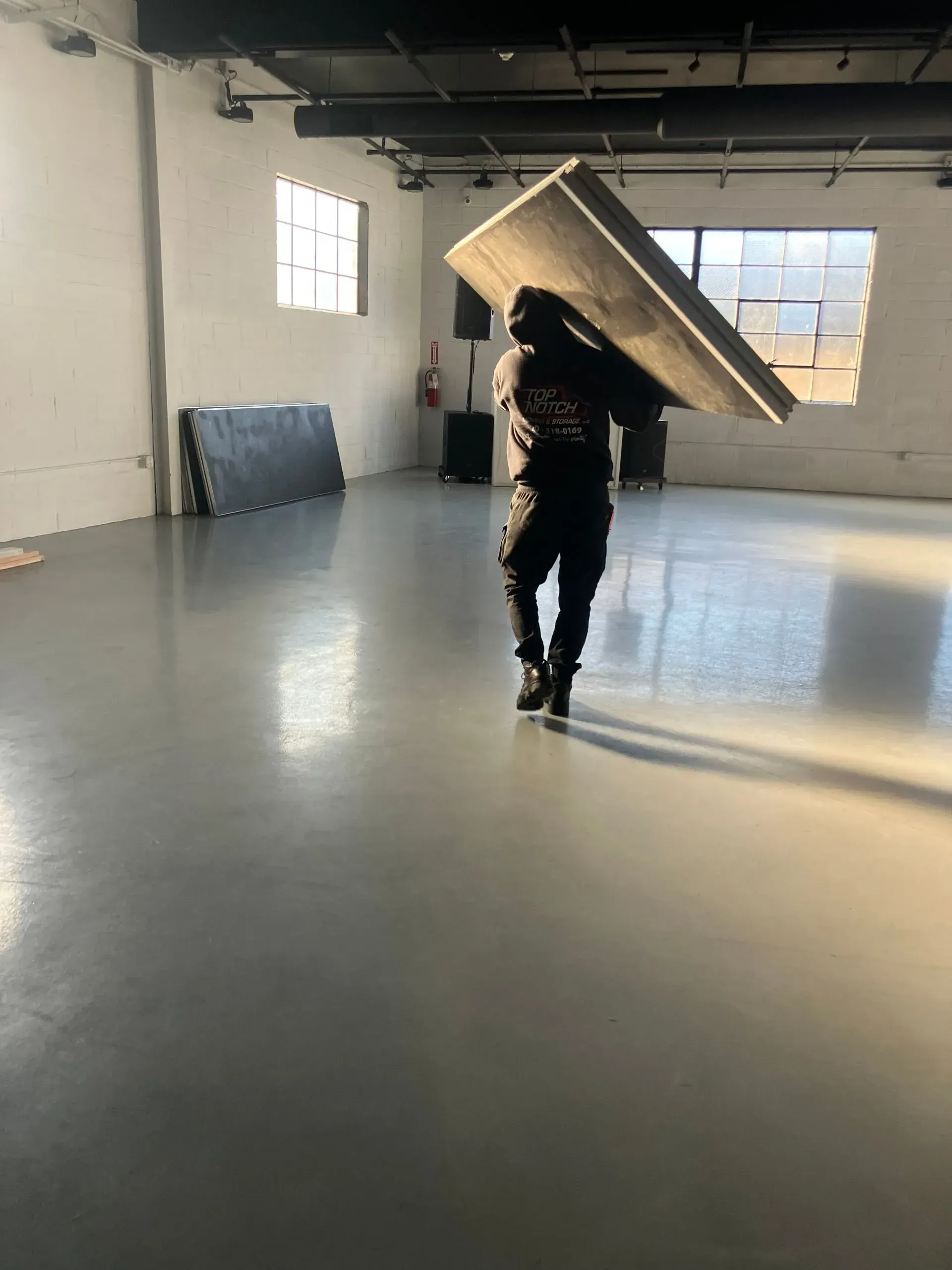Person carrying a large, dark rectangular object across a large, empty room with sunlight.