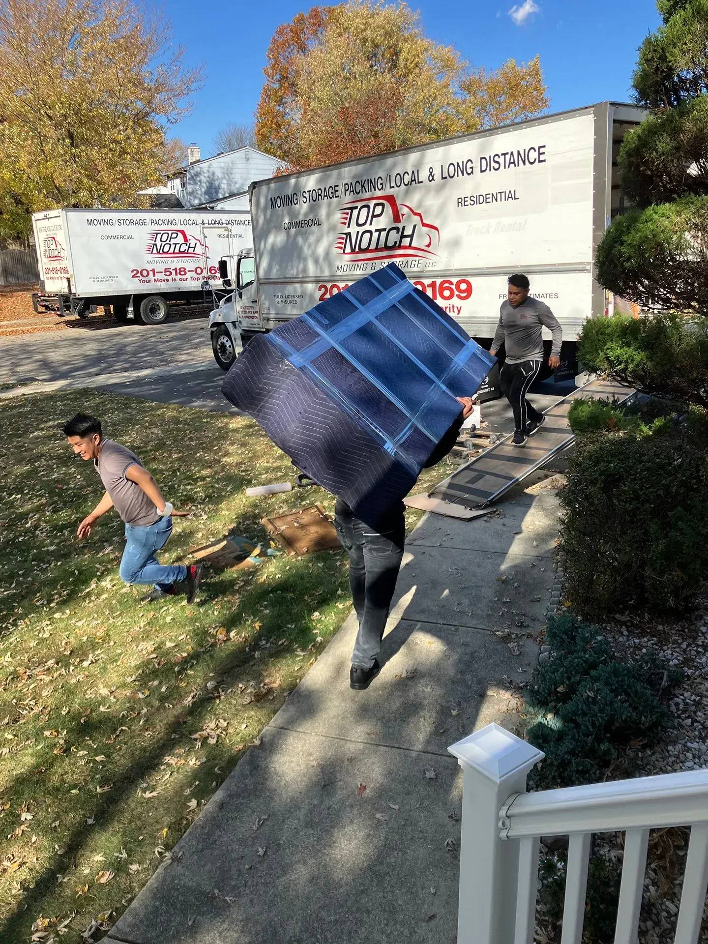 Three men carrying a large, dark solar panel. Moving trucks parked on a street near a house.