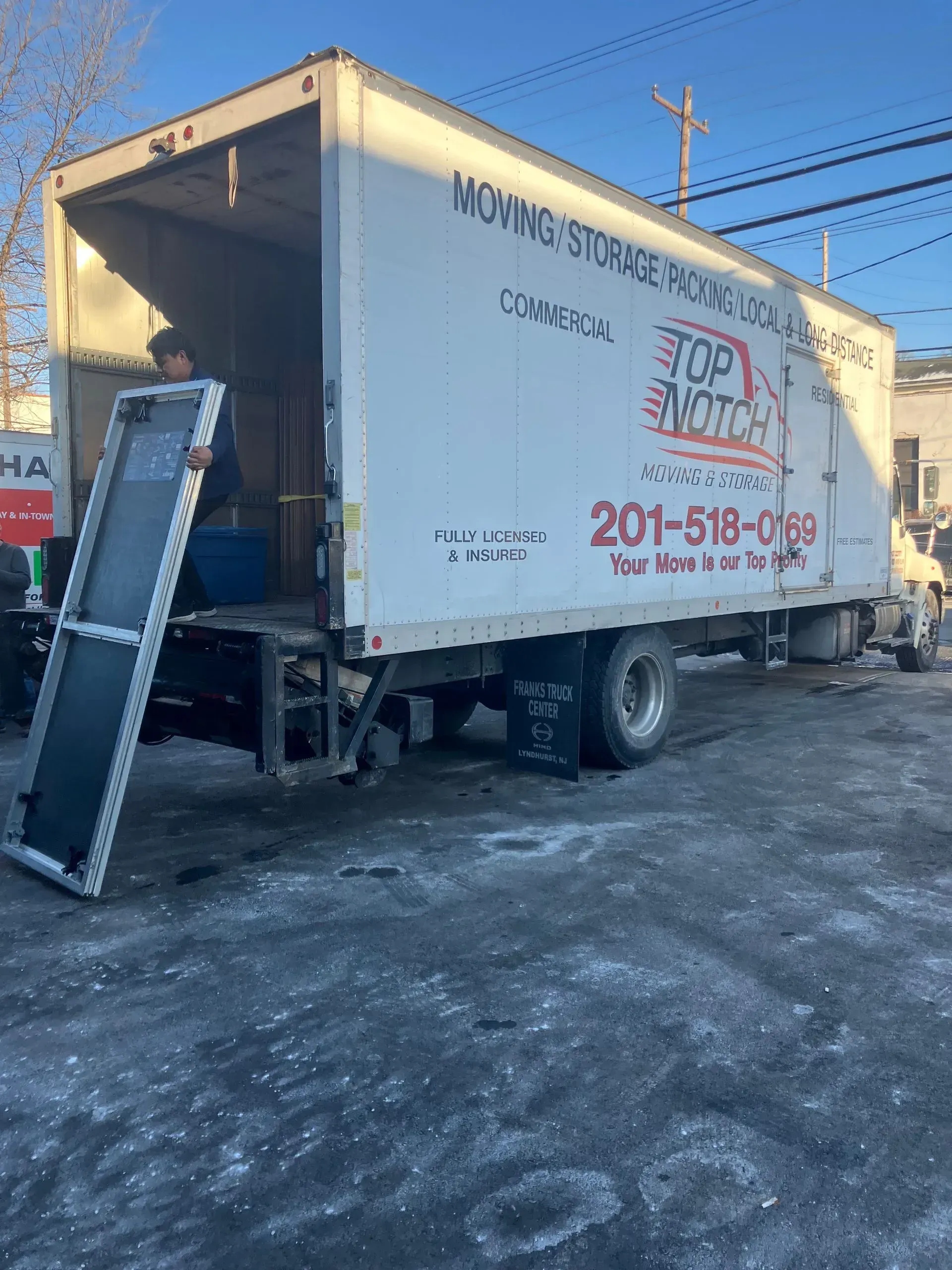Moving truck with ramp extended, parked on a snowy street, a person inside.