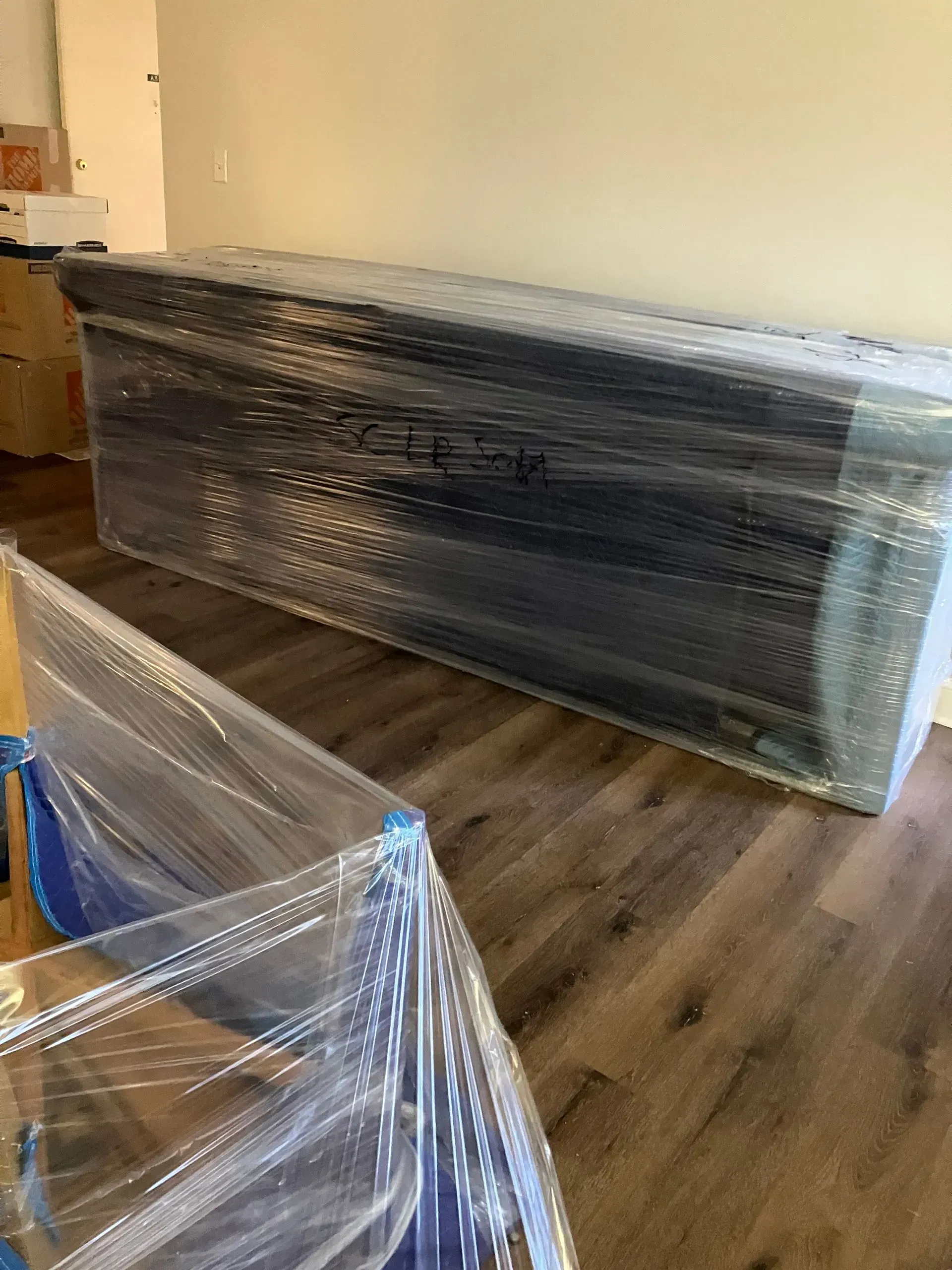 Furniture wrapped in clear plastic, presumably for moving.  Inside a room with wood floors and white walls.