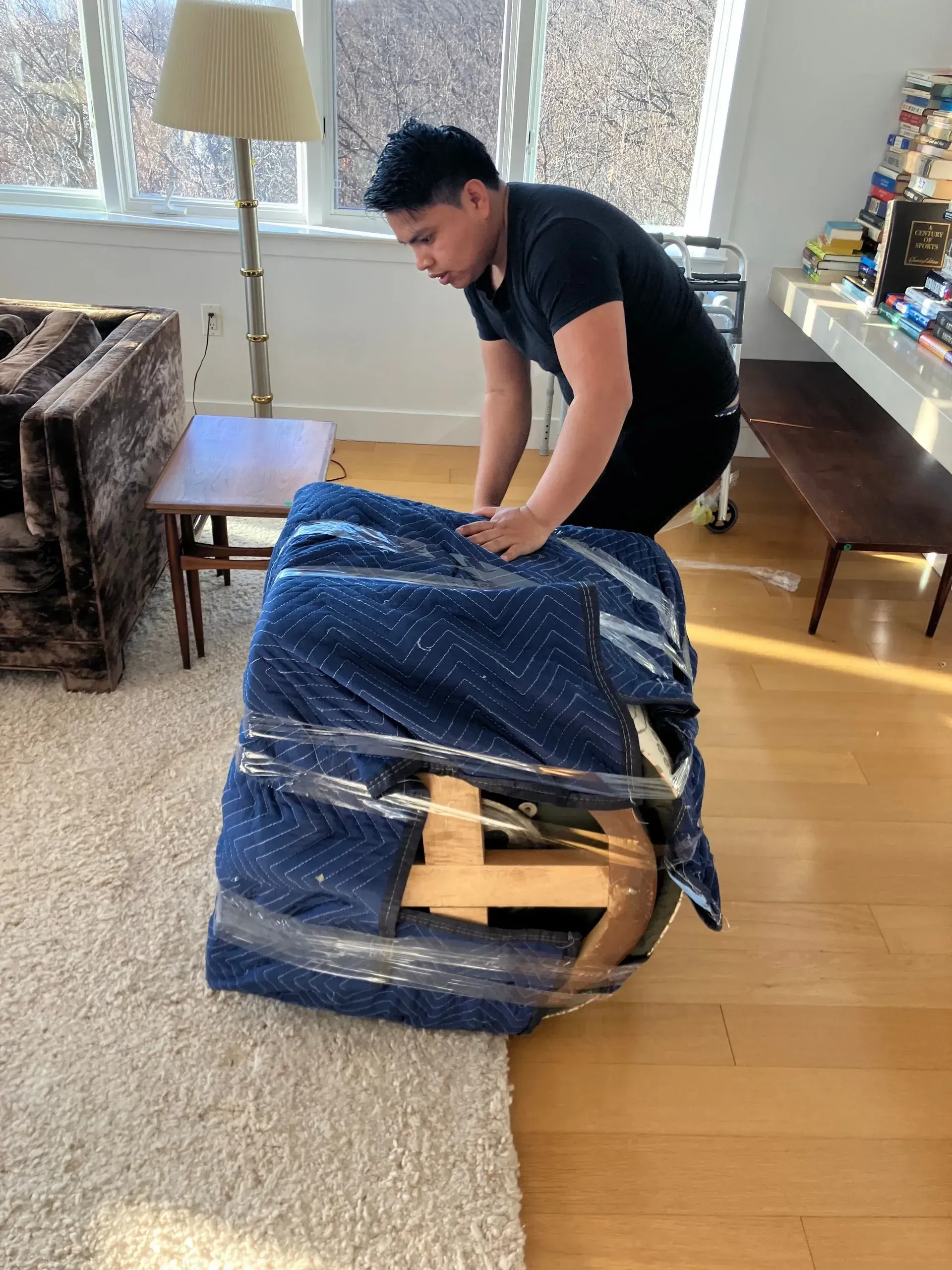 Person wrapping furniture with blue padding and plastic wrap, indoors.