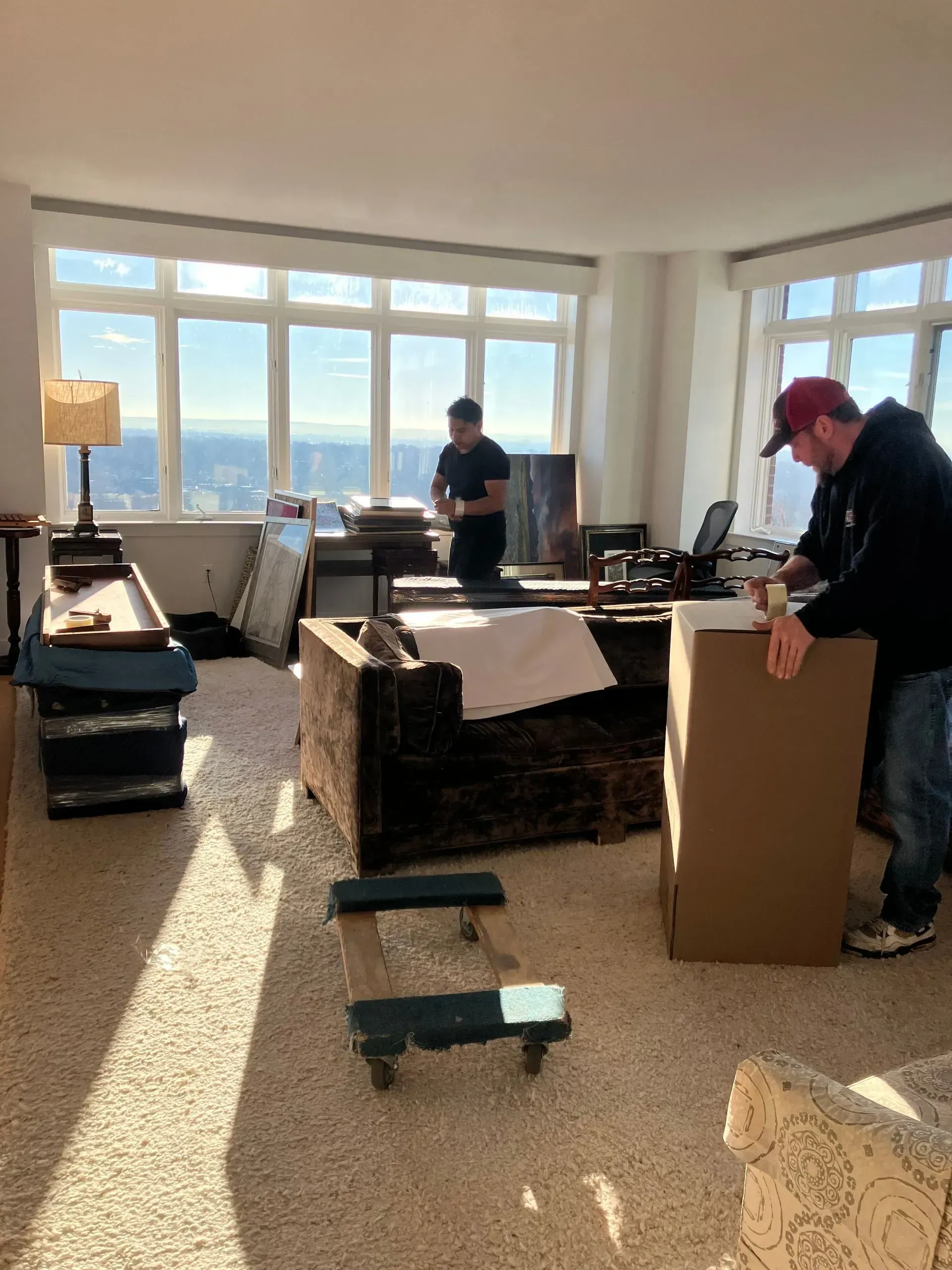 Two people moving furniture in a room with large windows, sunlight, and a city view.