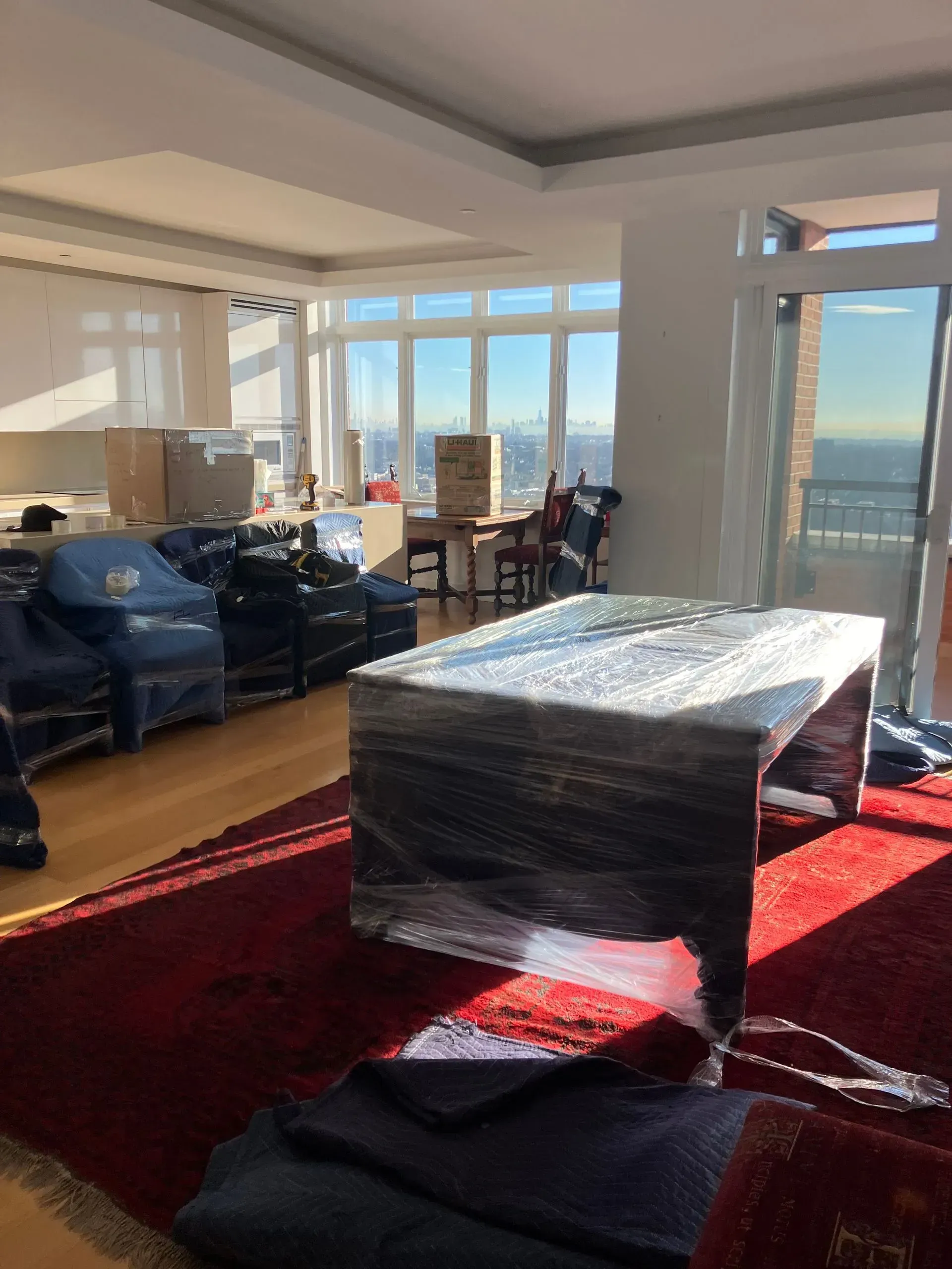 A room with furniture wrapped for moving, sunlight streaming in. Boxes and chairs are present.