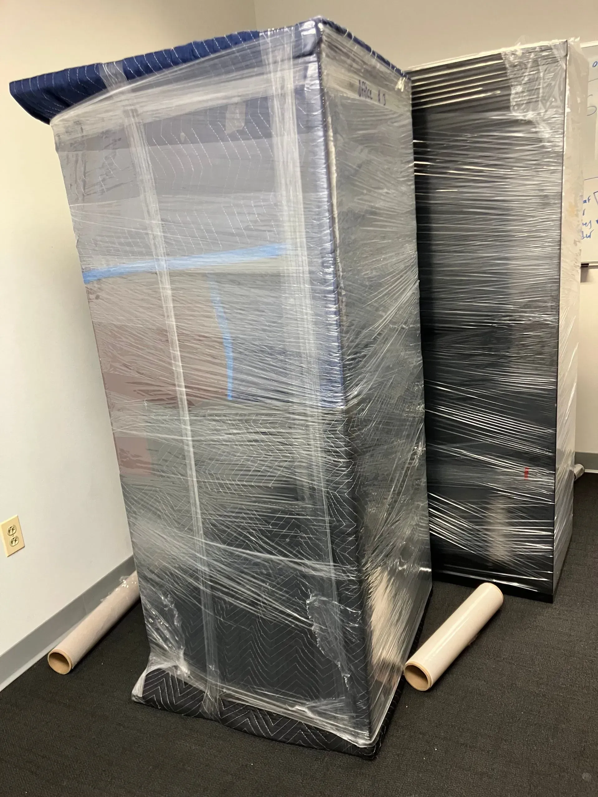 Two tall, wrapped black cabinets stand on a dark floor, rolls of wrapping nearby.