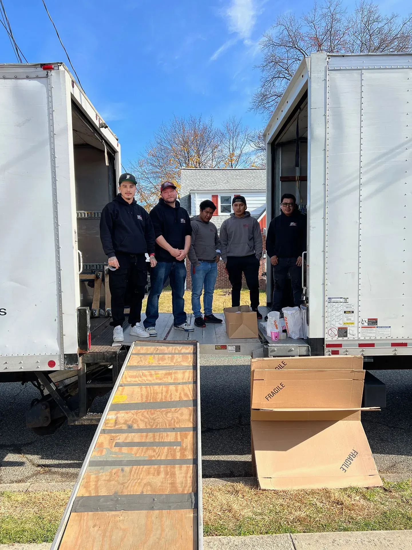 Five people stand near open moving trucks. One truck has a ramp. Boxes sit nearby.