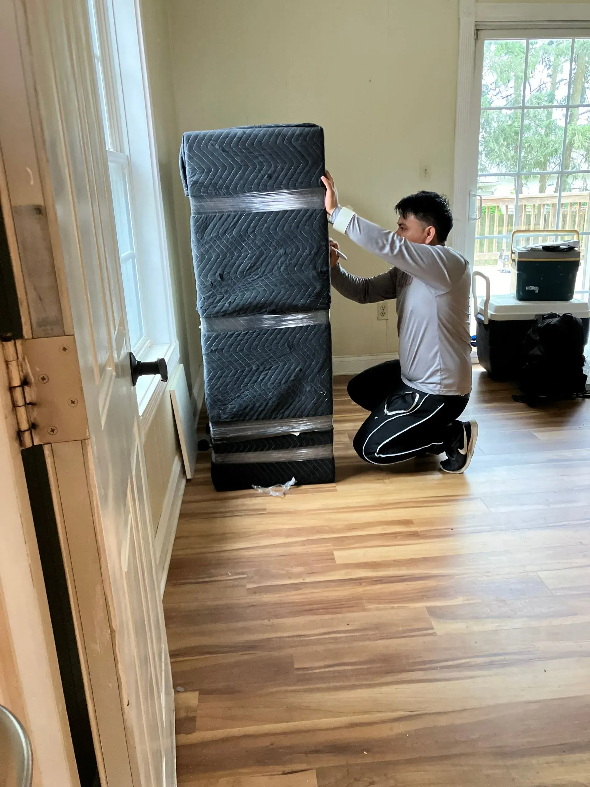 Person kneeling, securing a wrapped, tall object with tape in a room with hardwood floors and a window.