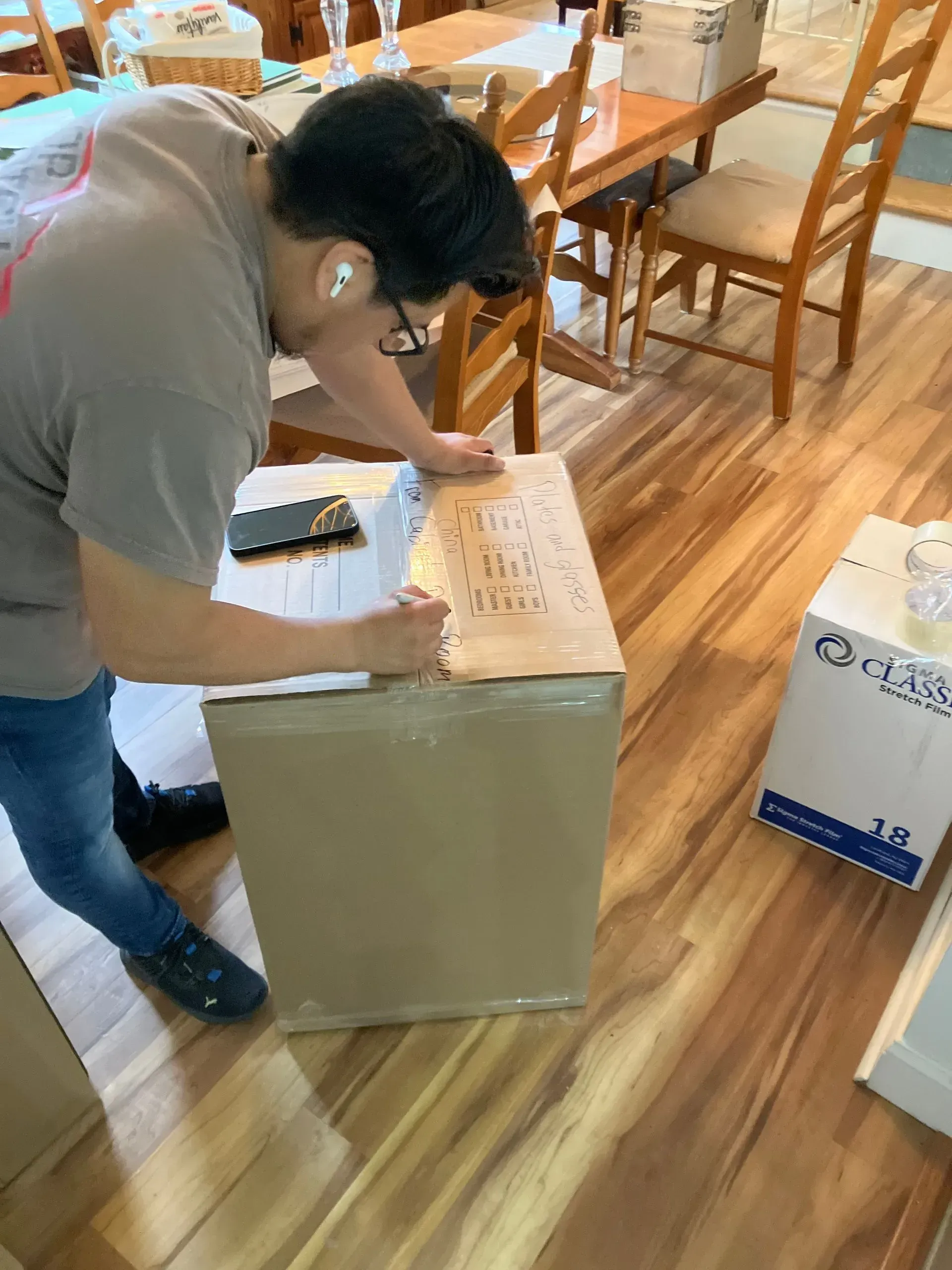 Person writing on a cardboard box, indoors, with wood flooring and furniture in the background.