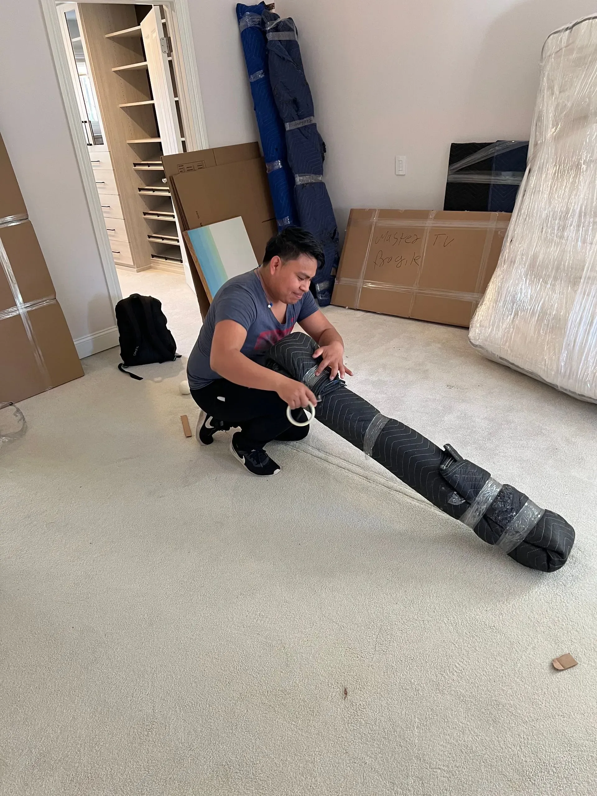 Person kneeling, wrapping a long, black object, possibly a rolled-up rug, in a room with boxes and a closet.