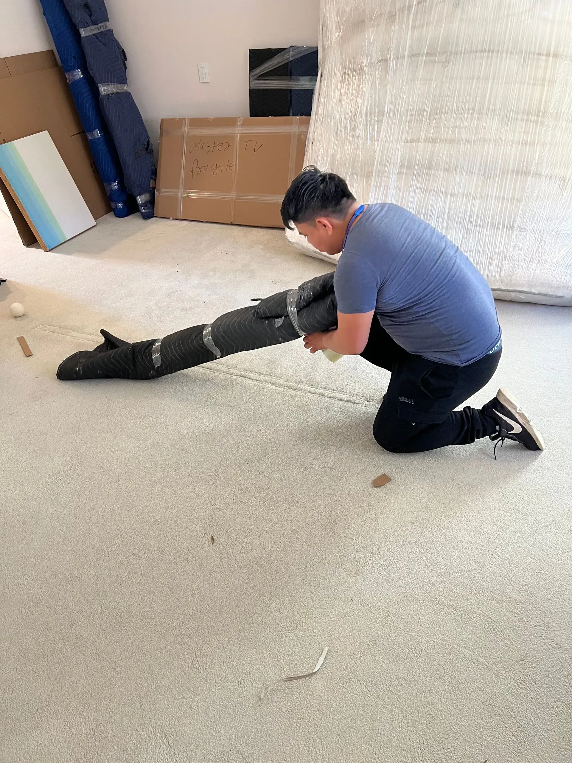 Man kneeling on carpet, rolling a wrapped object.  Blue shirt, dark pants, light-colored room.