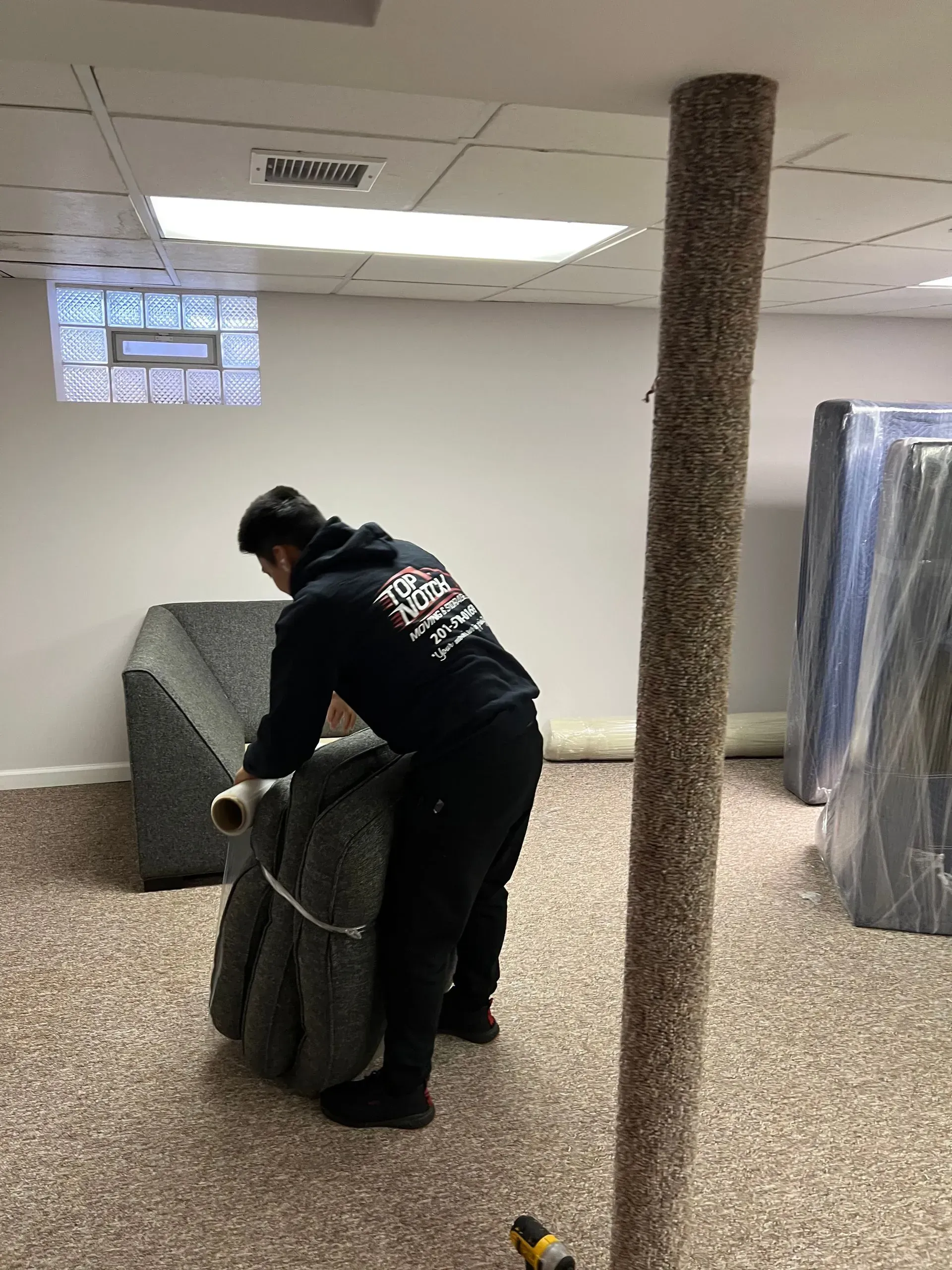 Person rolling a carpet in a room with a support column and furniture covered in plastic.