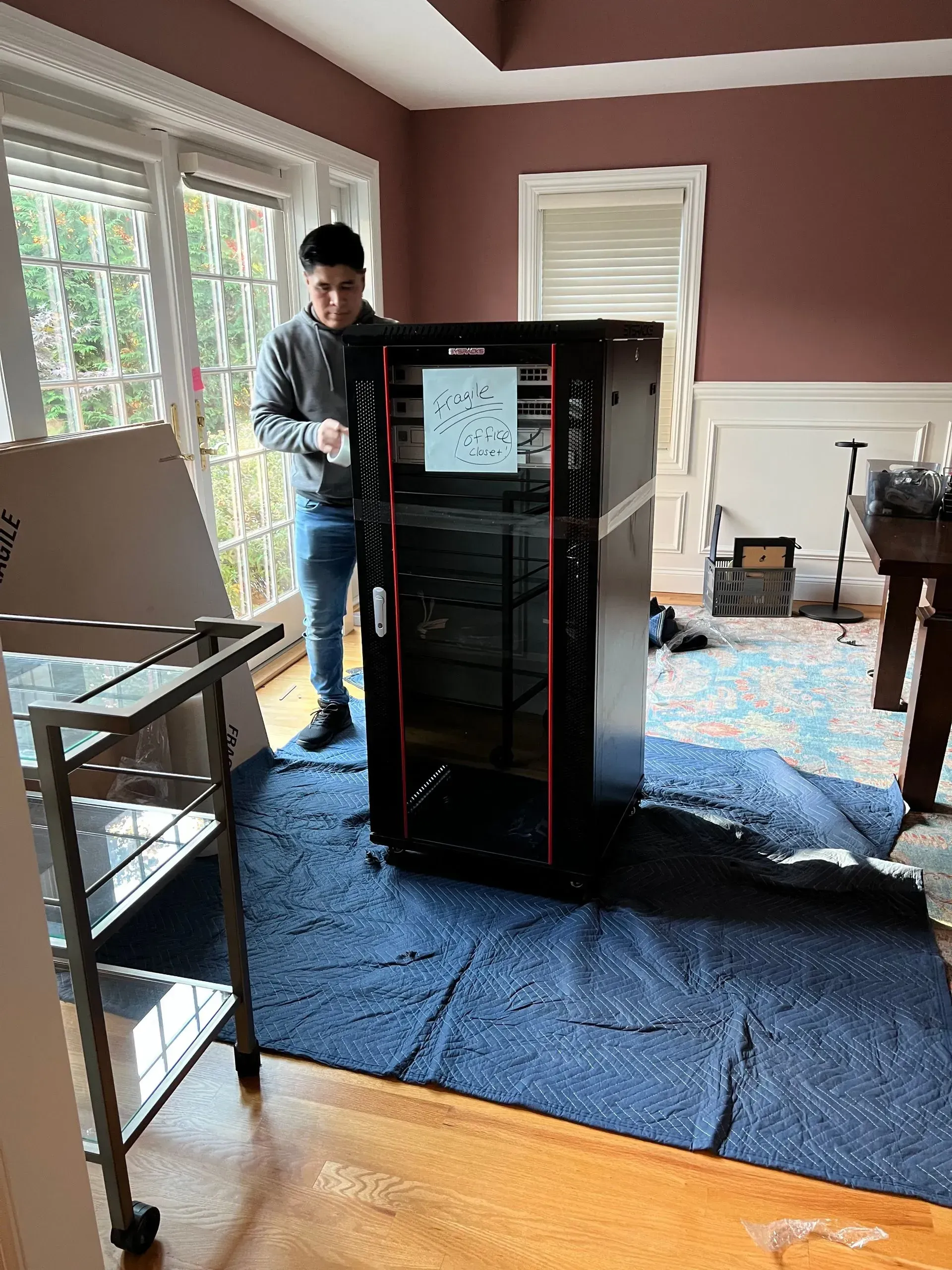 Man near a large server rack on a blue mat in a room with a window, pink wall, and a rolling cart.