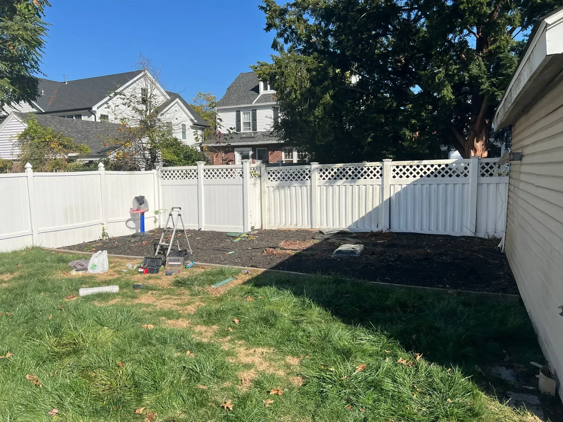 Backyard with white fence, patches of grass, and freshly mulched garden beds. Tools and containers are present.