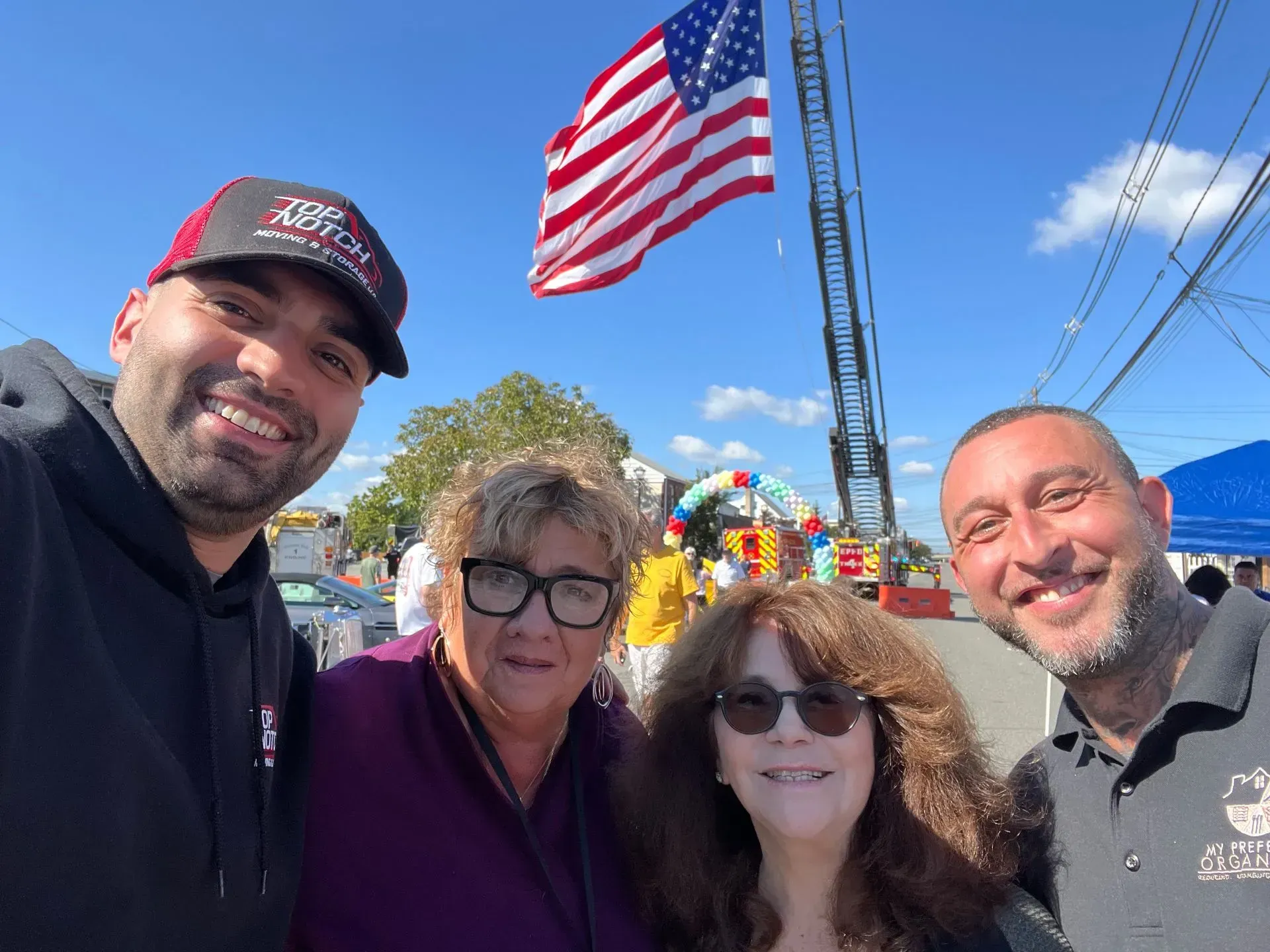 Four people smiling, posing for a photo outdoors. American flag and fire ladder in the background.