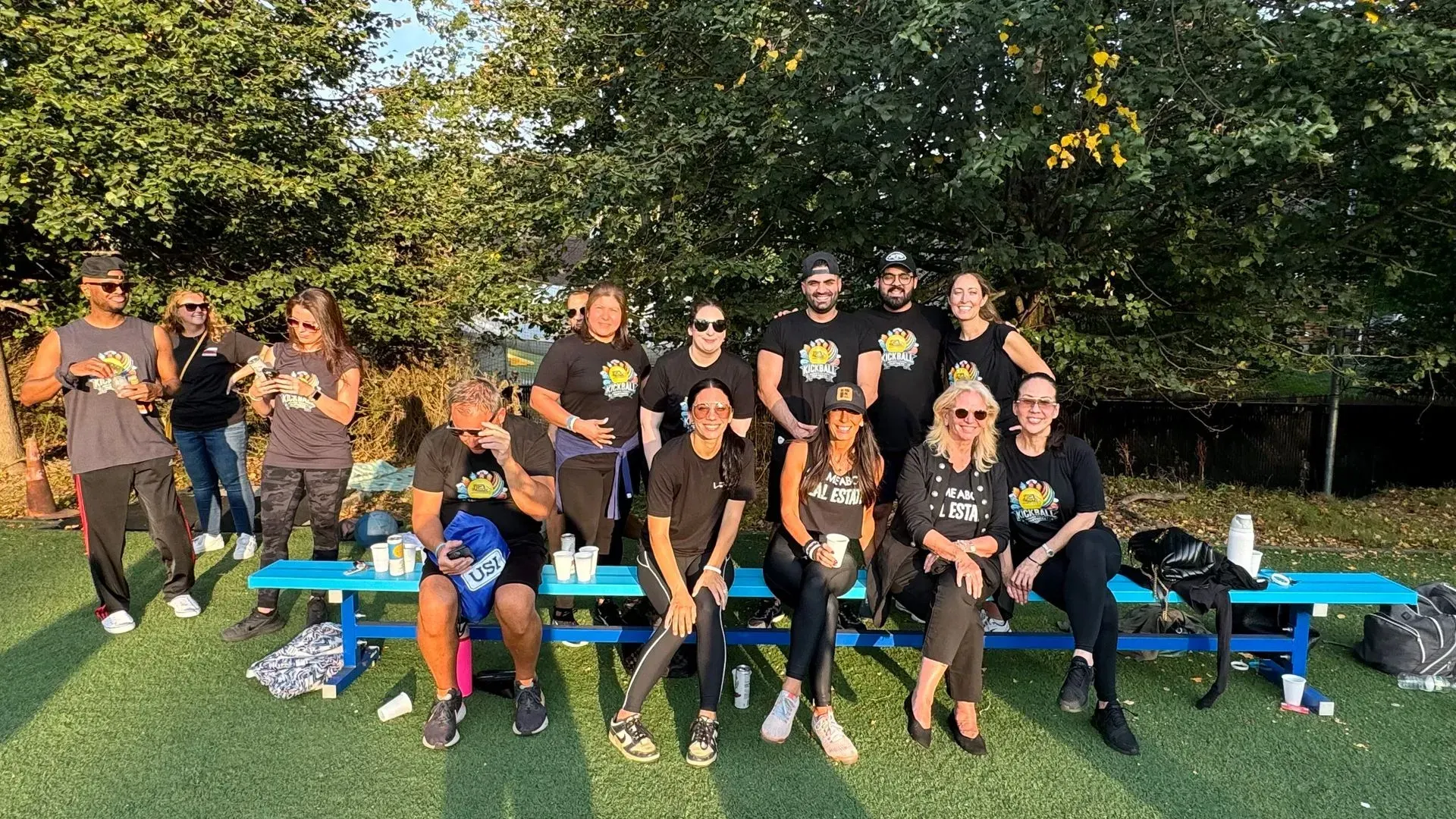 Group of people in black shirts and sunglasses, smiling outdoors. Some seated on a blue bench on grass.