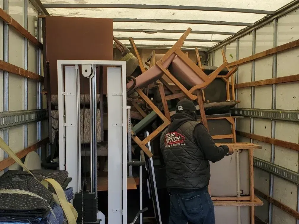 Man loads furniture into a moving truck. Brown and white furniture, the man wears a dark vest and jeans.