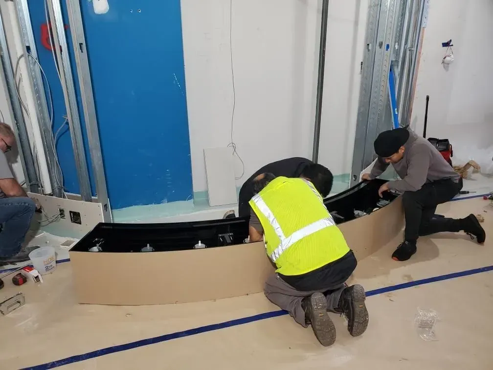 Three people installing a curved, black structure in a room under construction, one wearing a reflective vest.