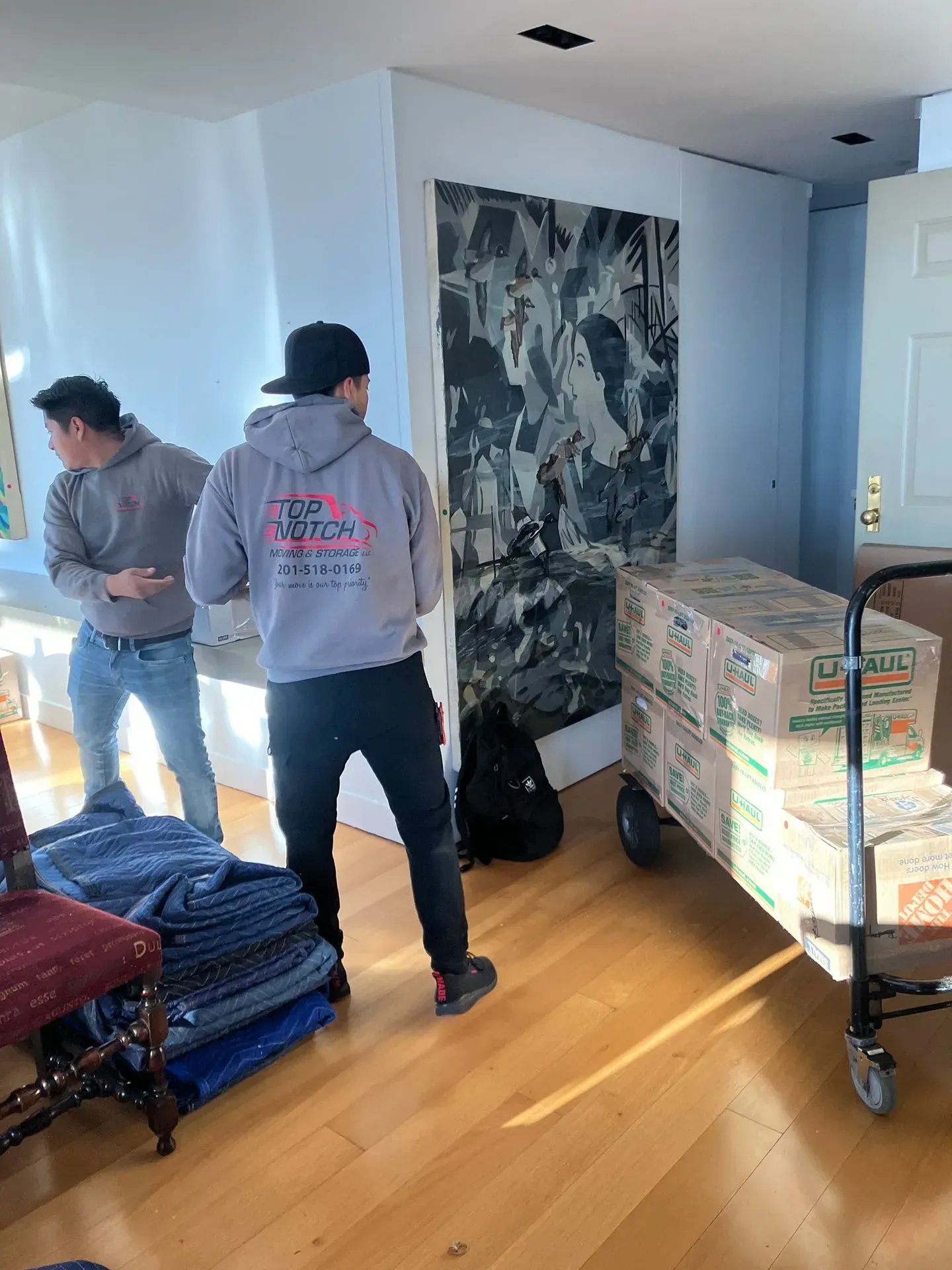 Two movers with boxes on a hand truck inside a home, near a large artwork.