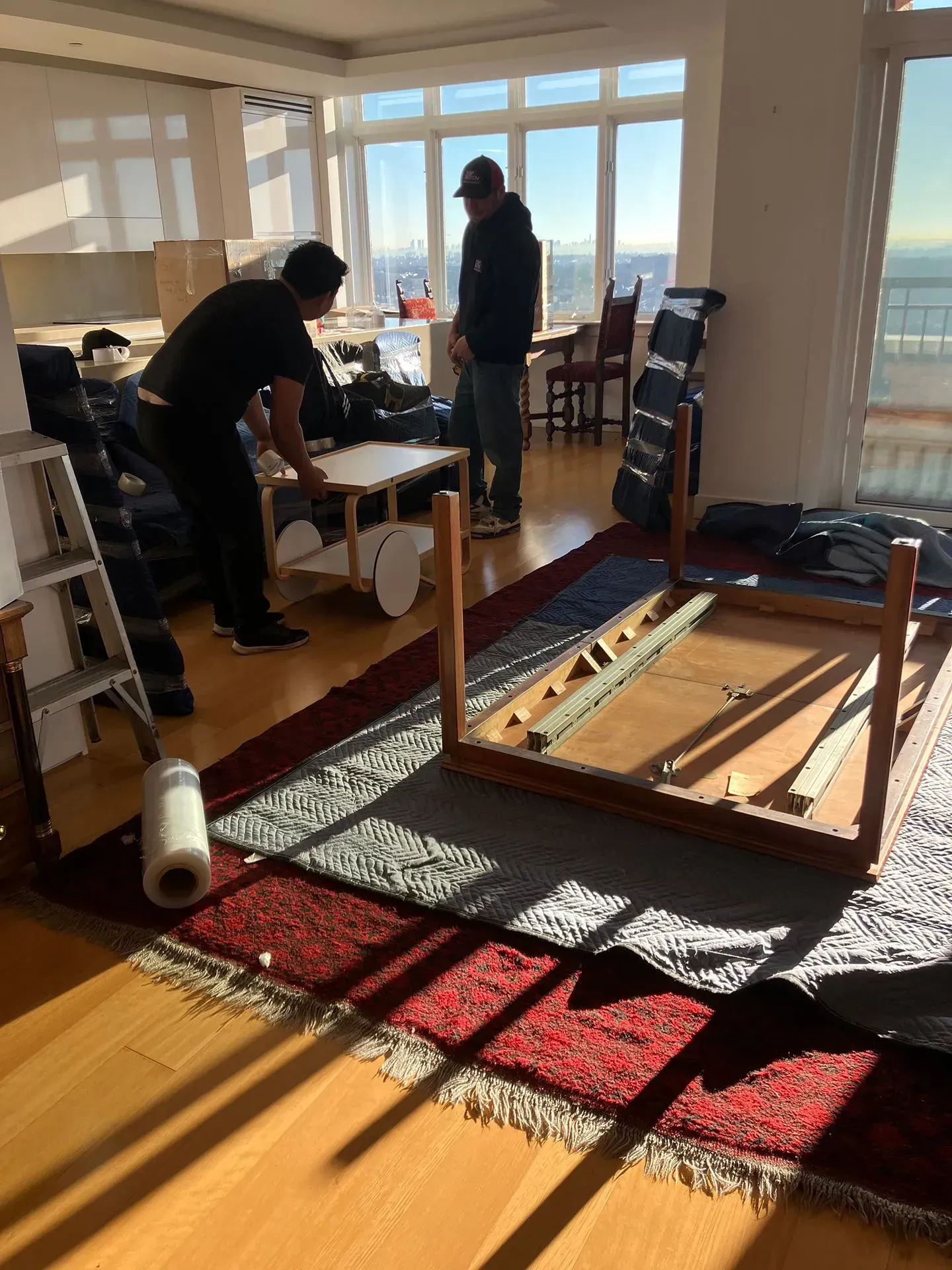 Two people assembling furniture in a bright room with city views. One is placing a tabletop, the other observes.