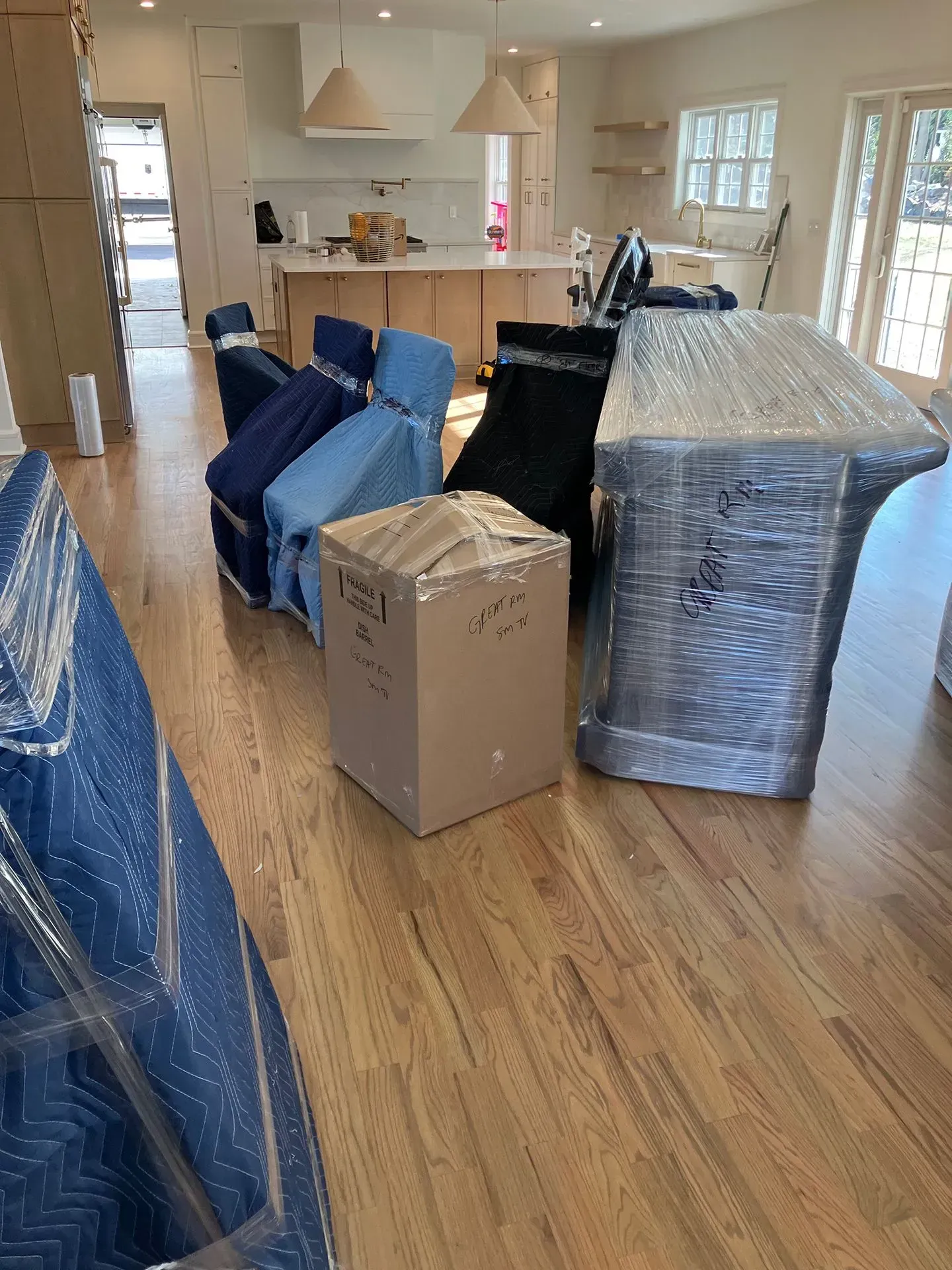 Furniture wrapped in plastic and moving blankets on hardwood floor in a home.