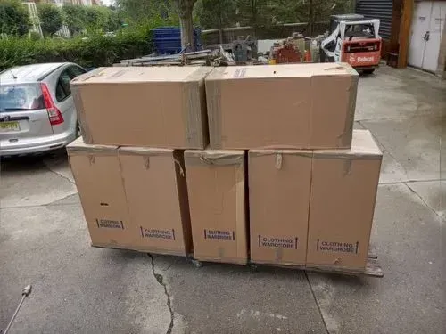 Cardboard boxes stacked outdoors on a concrete surface, near a car and equipment.